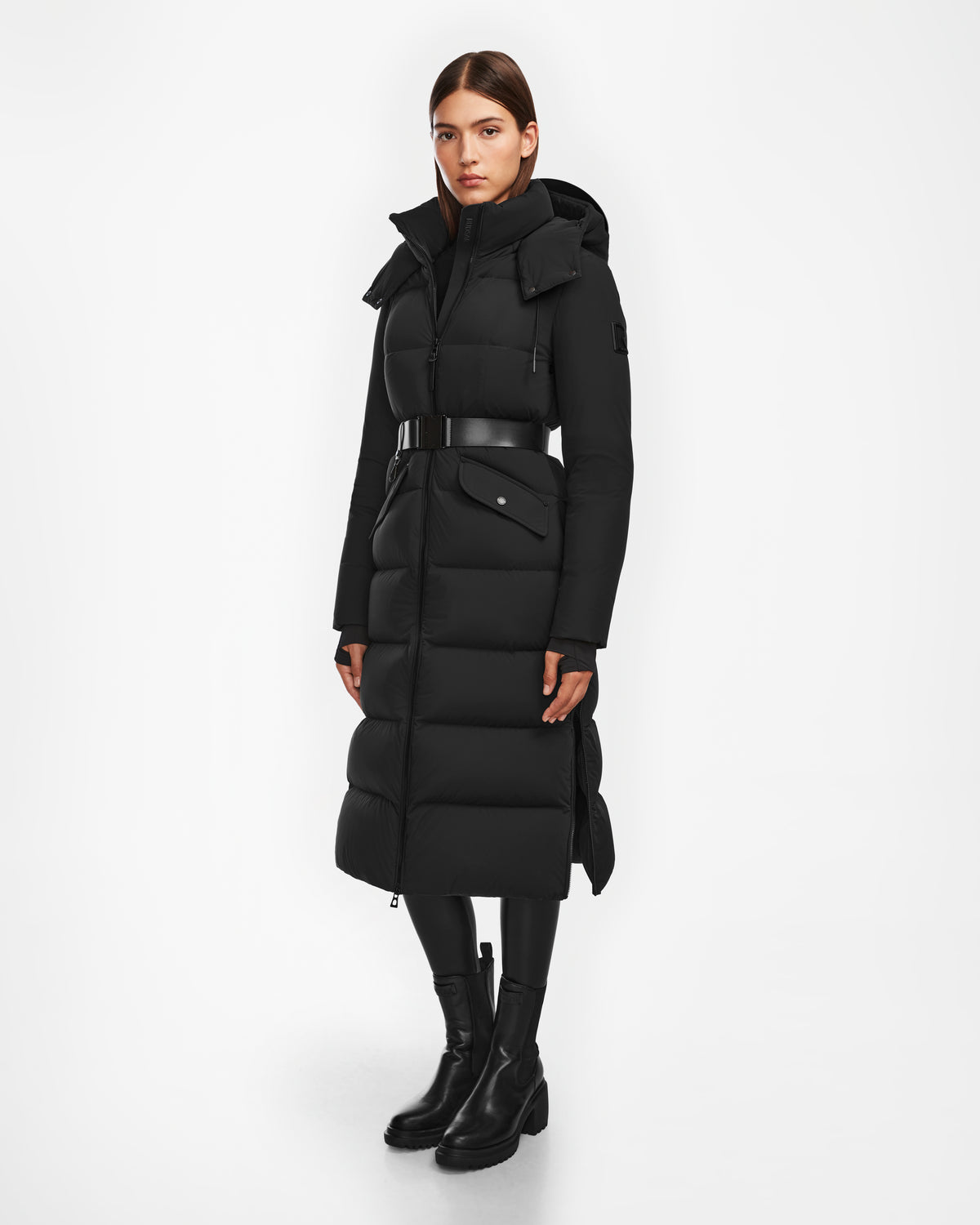 A woman wearing the Rudsak CALI - 8124600 Black long down puffer with belt, black leggings, and ankle boots stands against a white background; the jacket features a high collar and hood. She looks straight ahead with a neutral expression.