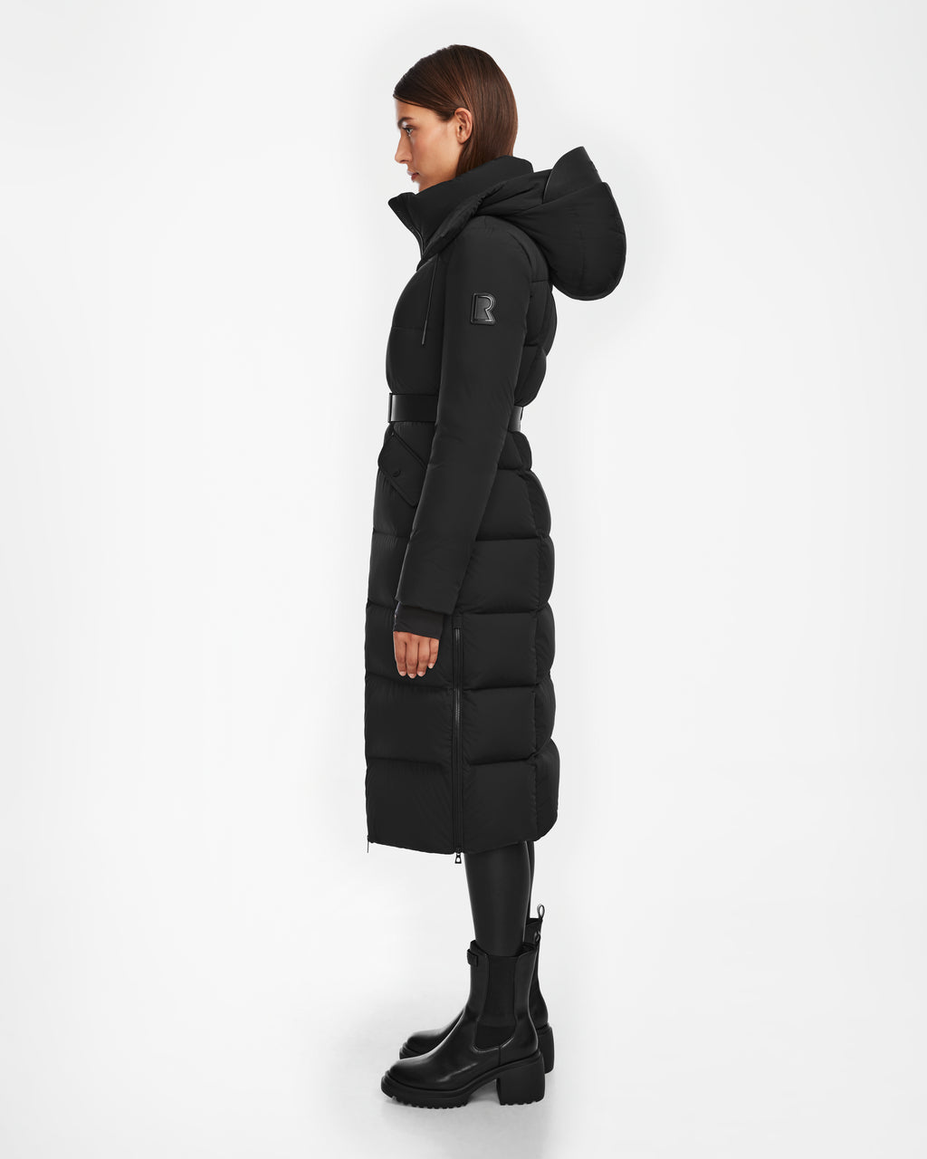 A woman in profile wears the Rudsak CALI - 8124600 Black women’s long down puffer jacket with a hood, black belt, and chunky black boots (RDS-certified), set against a plain white background.