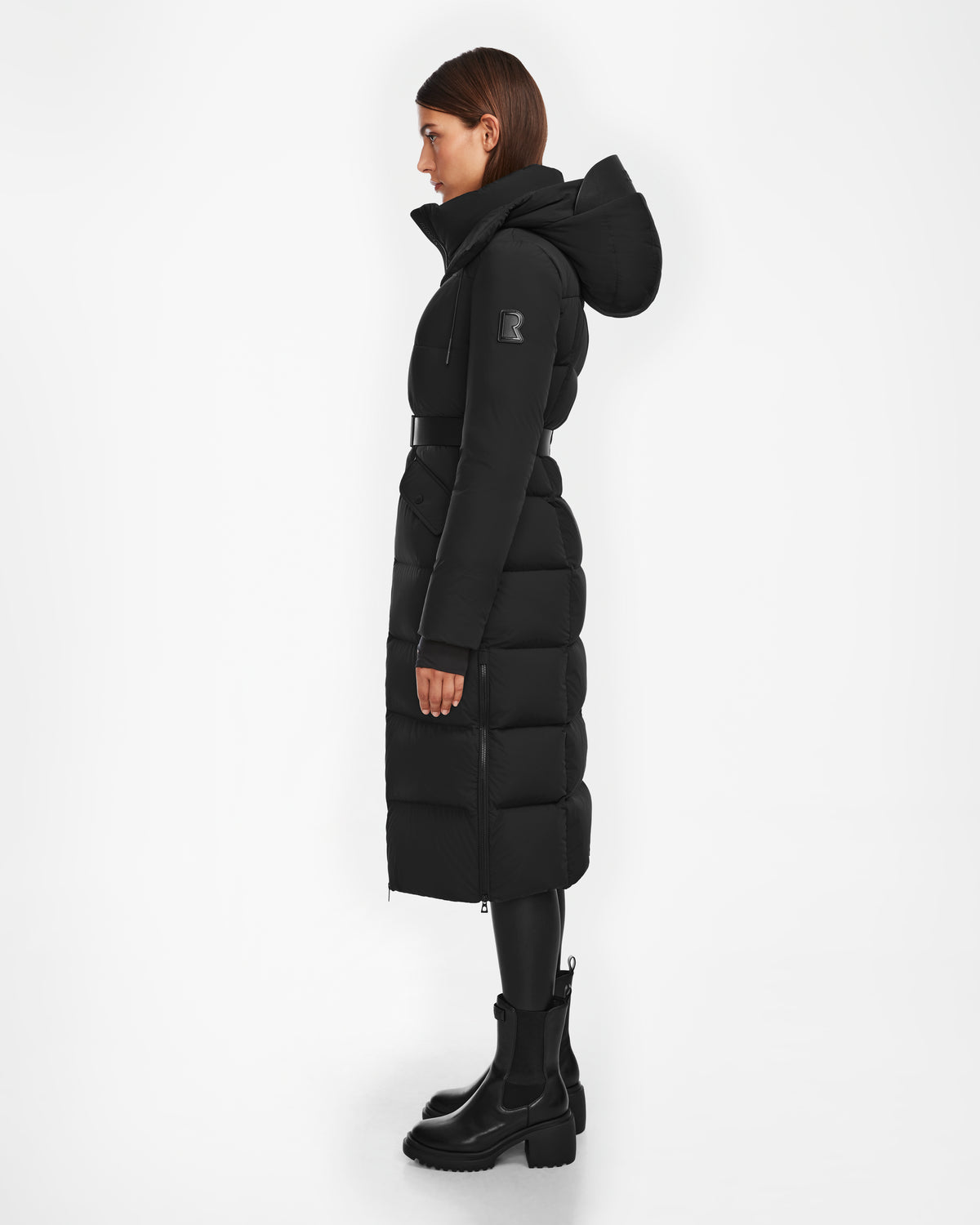 A woman in profile wears the Rudsak CALI - 8124600 Black women’s long down puffer jacket with a hood, black belt, and chunky black boots (RDS-certified), set against a plain white background.