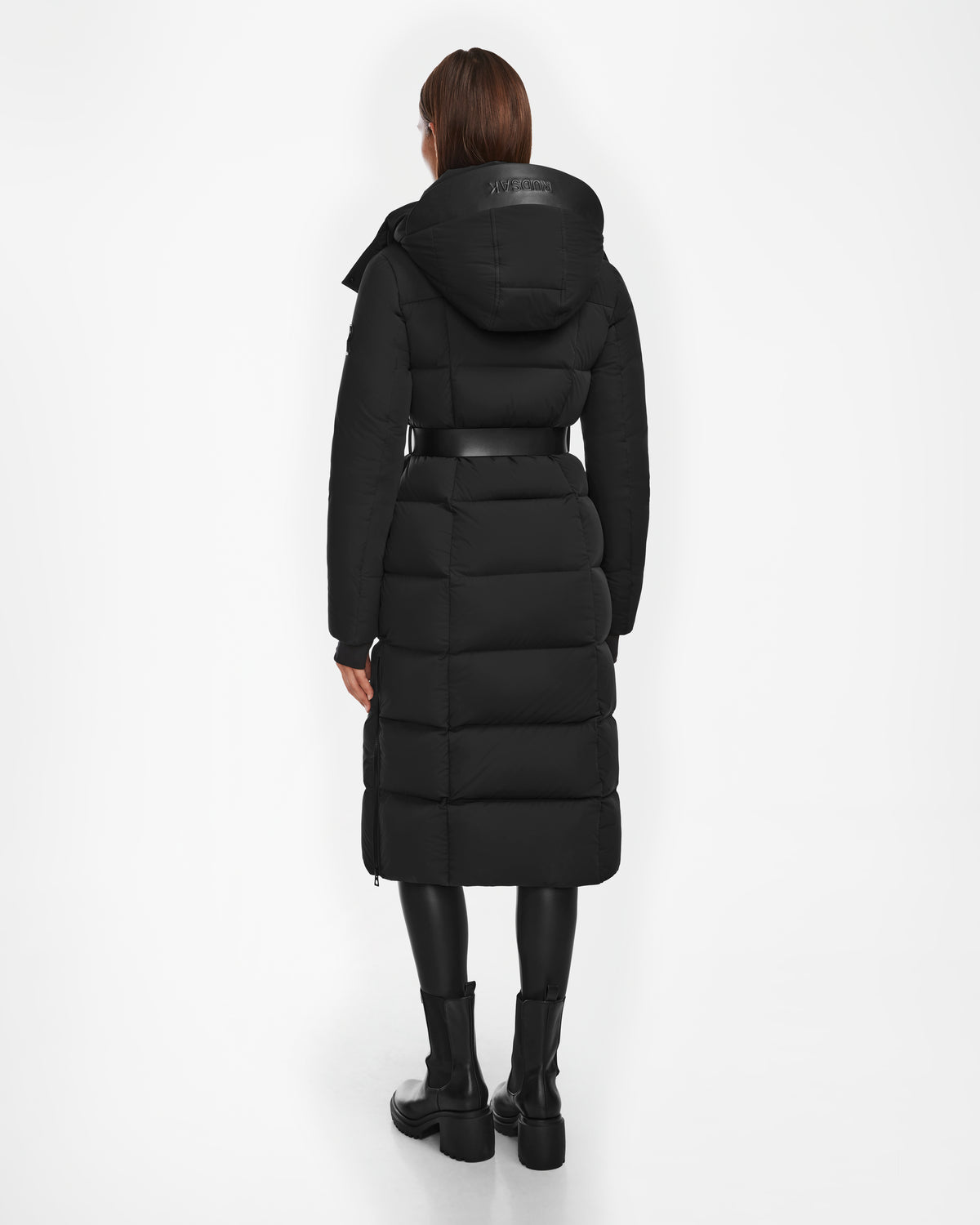 A person with straight brown hair wears the Rudsak CALI - 8124600 Black womens long down puffer with a hood, paired with black leggings and black boots, standing facing away on a white background.