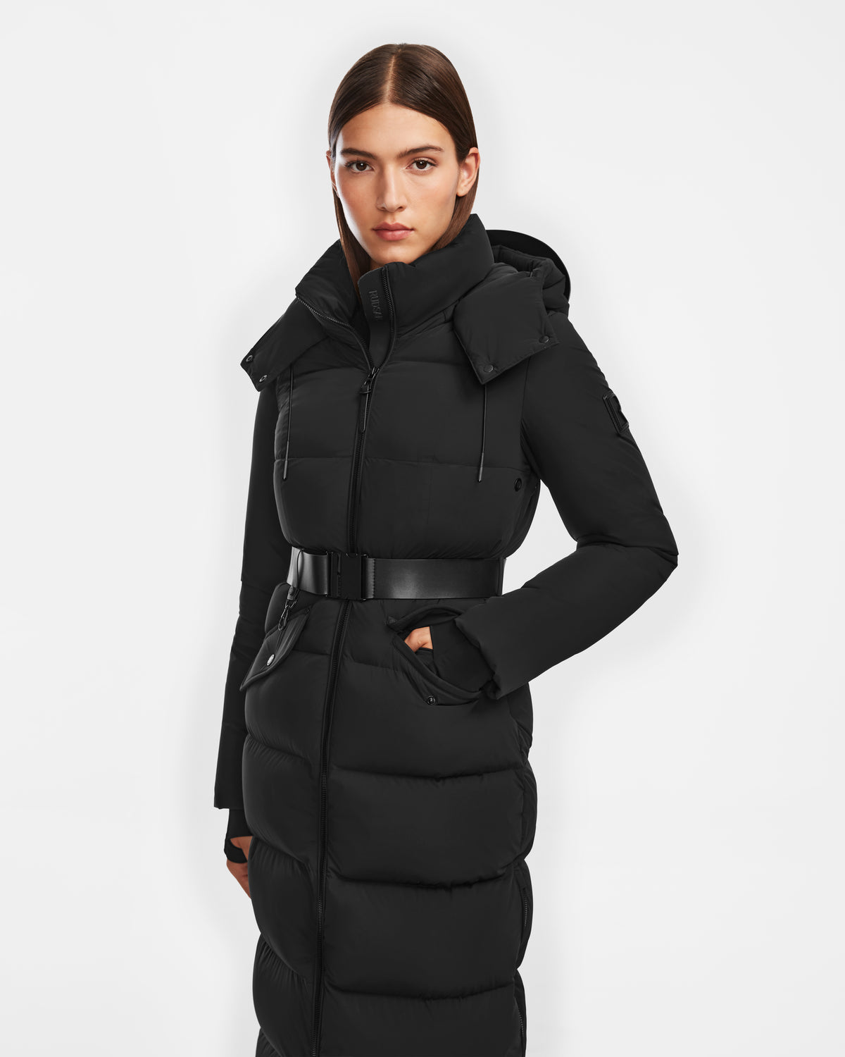 A woman with straight brown hair wears the Rudsak CALI - 8124600 Black long down puffer jacket with a hood and belt, standing against a white background and looking at the camera.