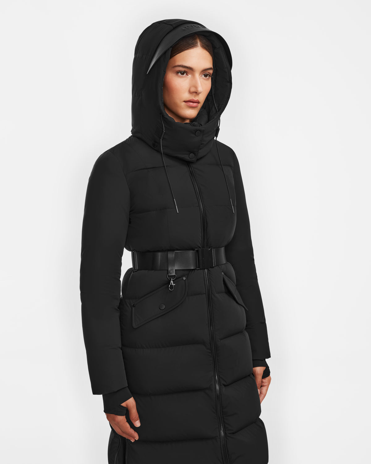 A woman wears the Rudsak CALI - 8124600 Black womens long down puffer jacket with hood and belt, made from RDS-certified down, standing against a plain white background.