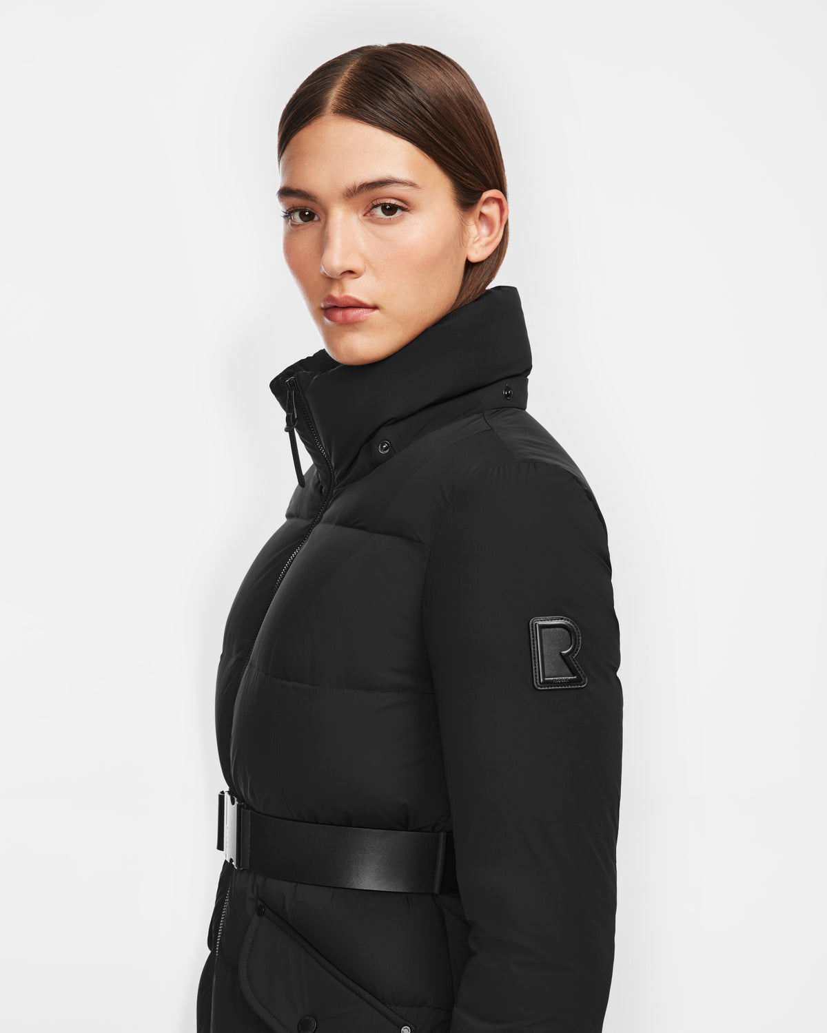 A woman with straight brown hair wears the Rudsak CALI - 8124600 Black long down puffer with a high collar, belt, and an R patch on the sleeve, featuring RDS-certified down. She stands against a white background.