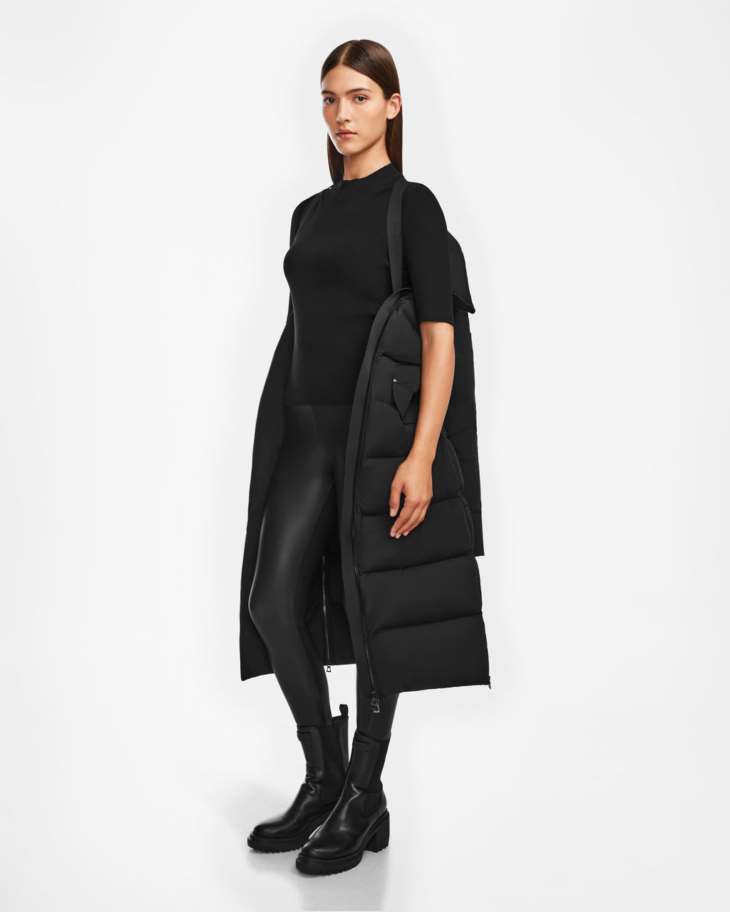 A woman stands slightly turned, wearing Rudsaks CALI - 8124600 Black womens long down puffer draped over her shoulders, paired with a black short-sleeve top, leggings, and mid-calf boots against a plain white background.