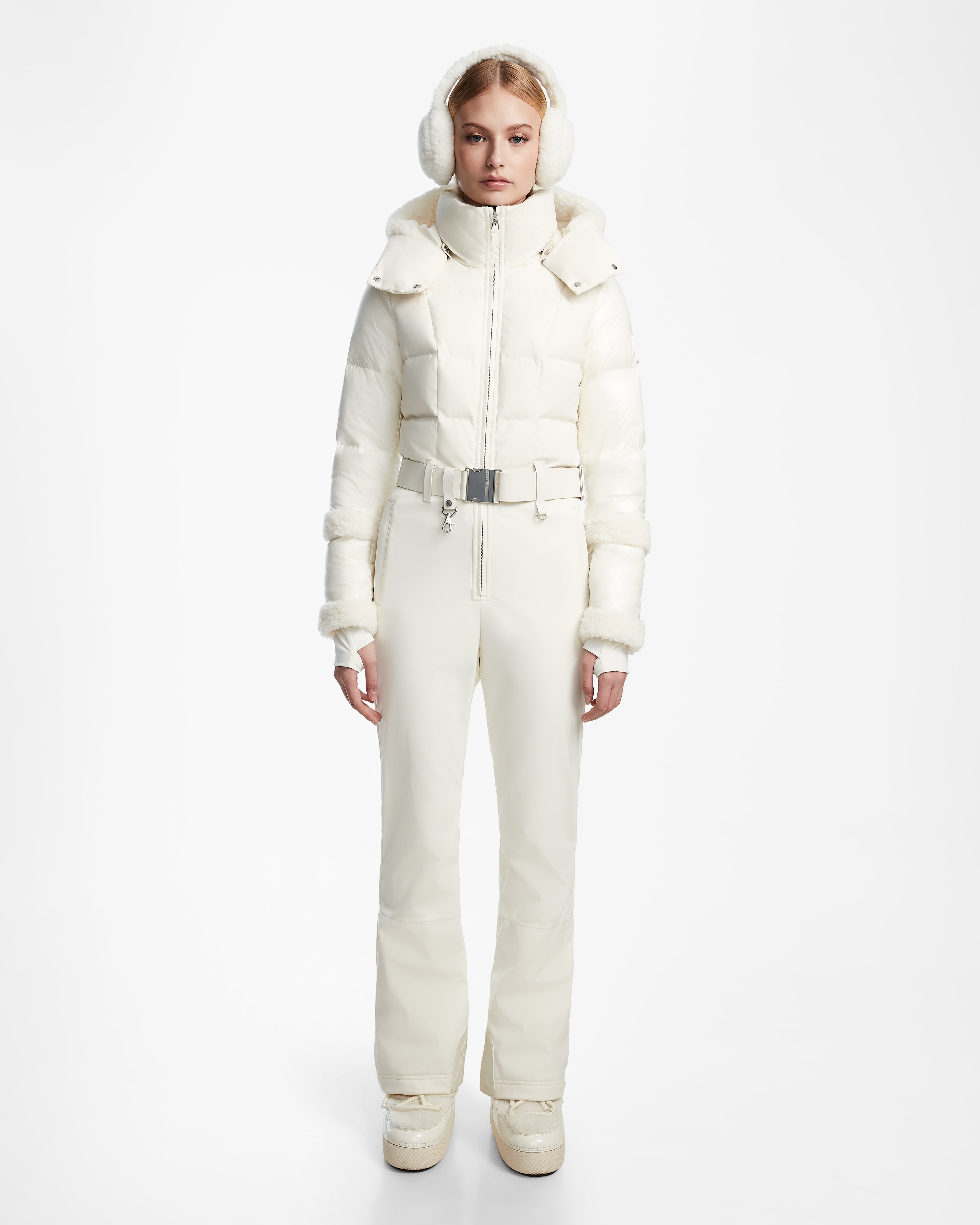 A woman stands facing forward against a white background wearing the Rudsak CARMEN - 8124783 Iceland white one-piece ski suit with RECCO rescue technology, paired with matching earmuffs and boots.