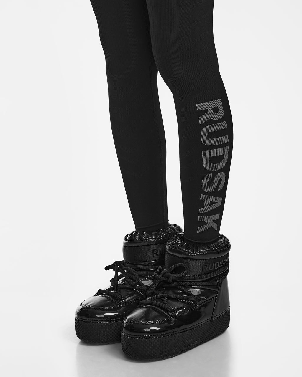 Person wearing black CARRIE - 8524619 high-top Rudsak boots and black ski underlayer leggings with RUDSAK vertically on the side, shown from the knees down on a white background.