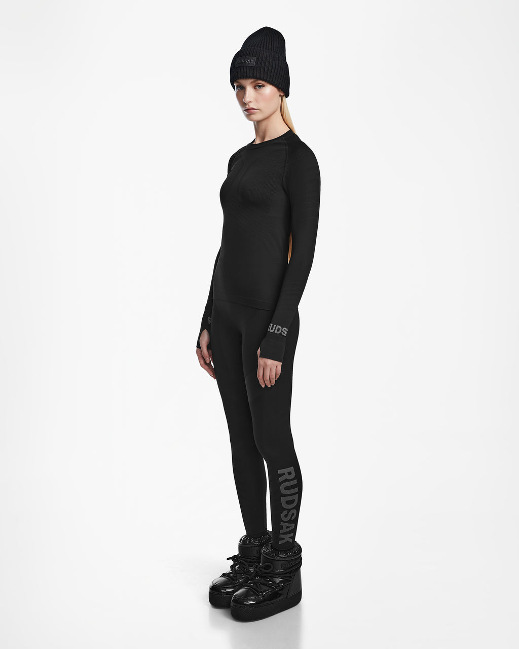 A woman with long blonde hair and a neutral expression stands against a white background wearing the Rudsak CARRIE - 8524619 Black ski underlayer top, matching black leggings with RUDSAK branding, boots, and a black beanie.