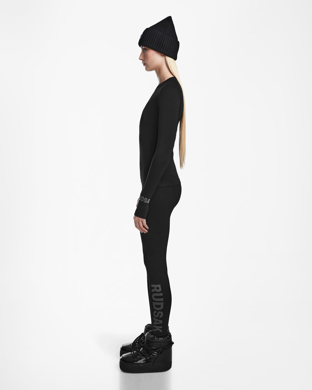 A woman in a black beanie, long-sleeve top, and Rudsak CARRIE - 8524619 Black leggings stands in profile against a white background, showcasing thermal performance and an elevated ski underlayer look completed with platform boots.