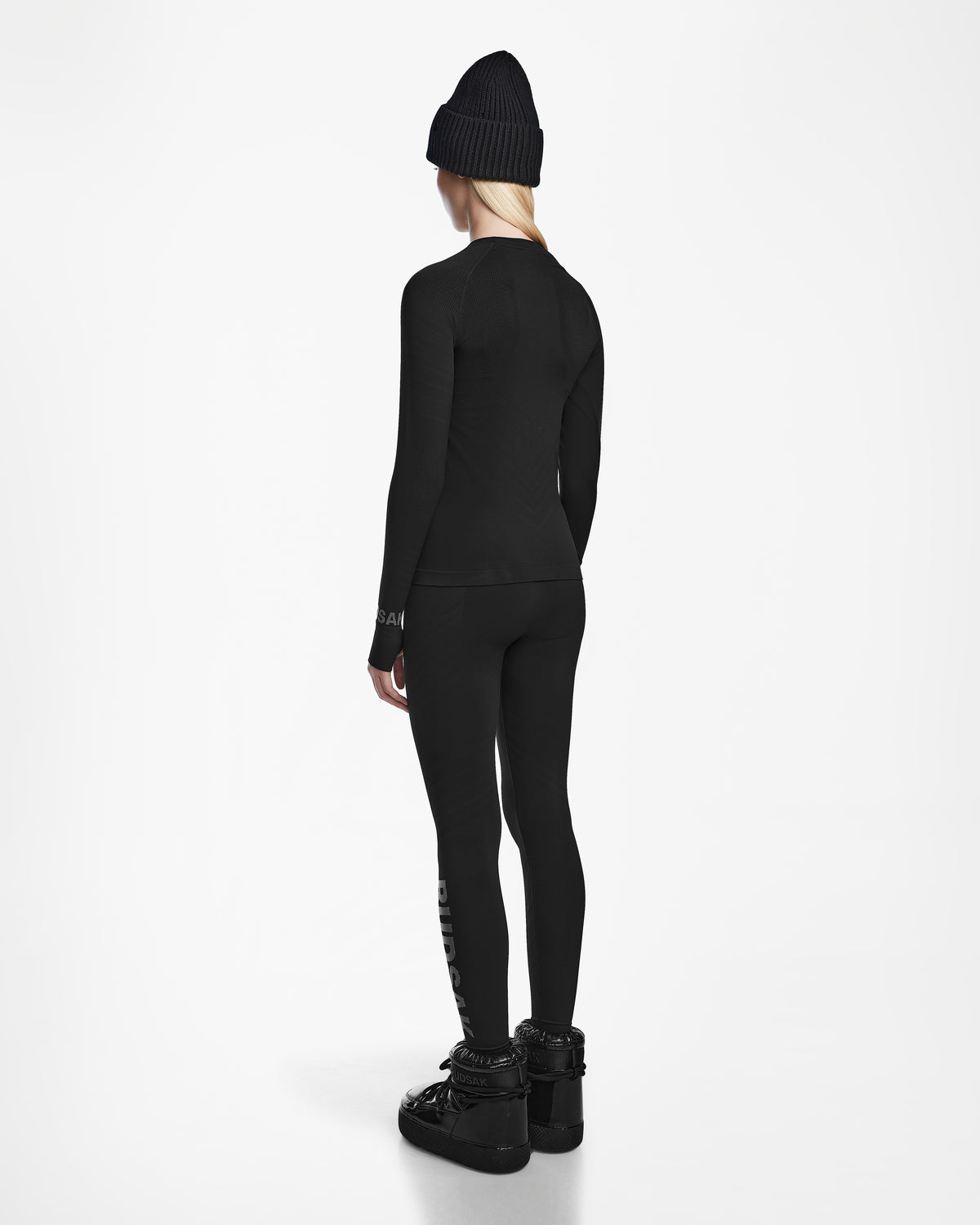 A person faces away, wearing the Rudsak CARRIE - 8524619 Black women’s stretch set with a black beanie and boots, set against a plain white background.