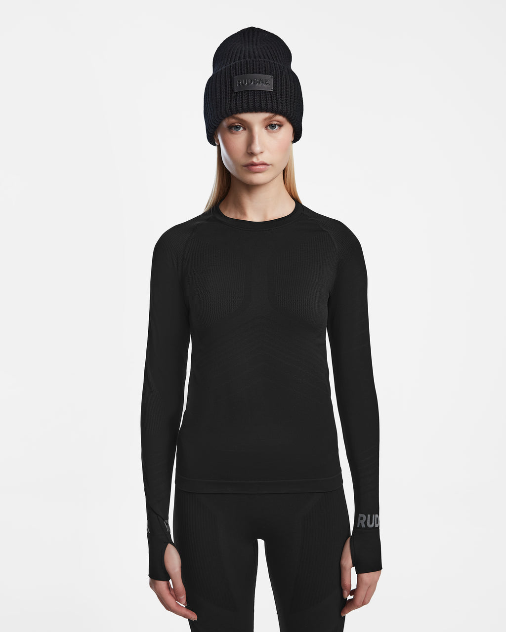 A woman wears the Rudsak CARRIE - 8524619 Black stretch base layer long-sleeve shirt, paired with black pants and a black knit beanie, standing facing forward against a plain white background.