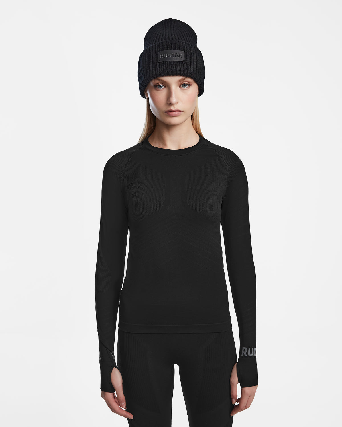 A woman wears the Rudsak CARRIE - 8524619 Black stretch base layer long-sleeve shirt, paired with black pants and a black knit beanie, standing facing forward against a plain white background.