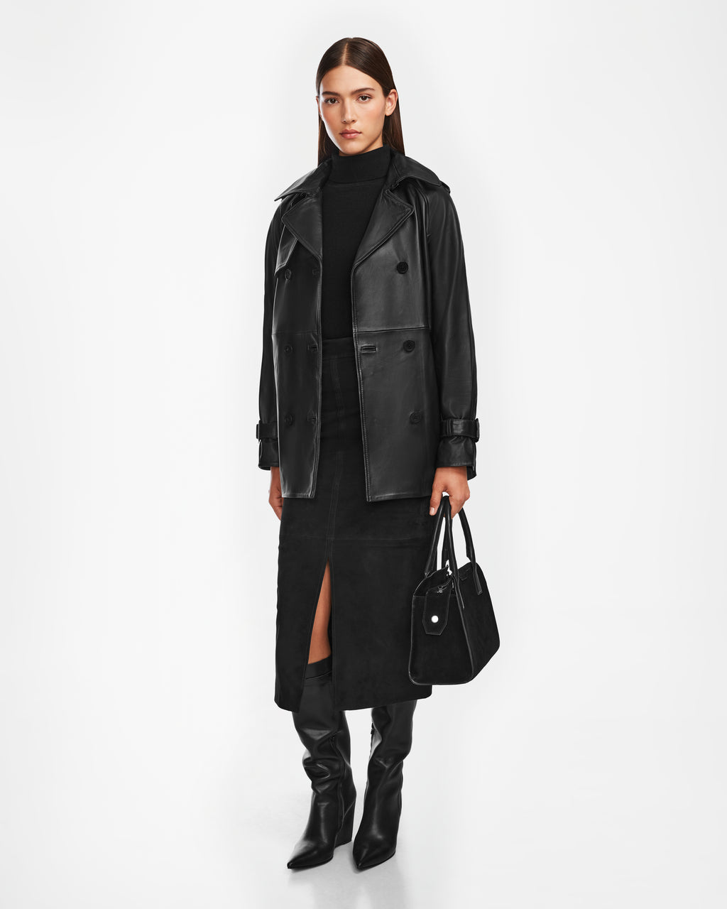 A woman stands against a white background wearing the Rudsak CASSIE M - 8124580 Black mid-length leather jacket, paired with a black turtleneck, front-slit skirt, knee-high boots, and holding a chic black handbag.