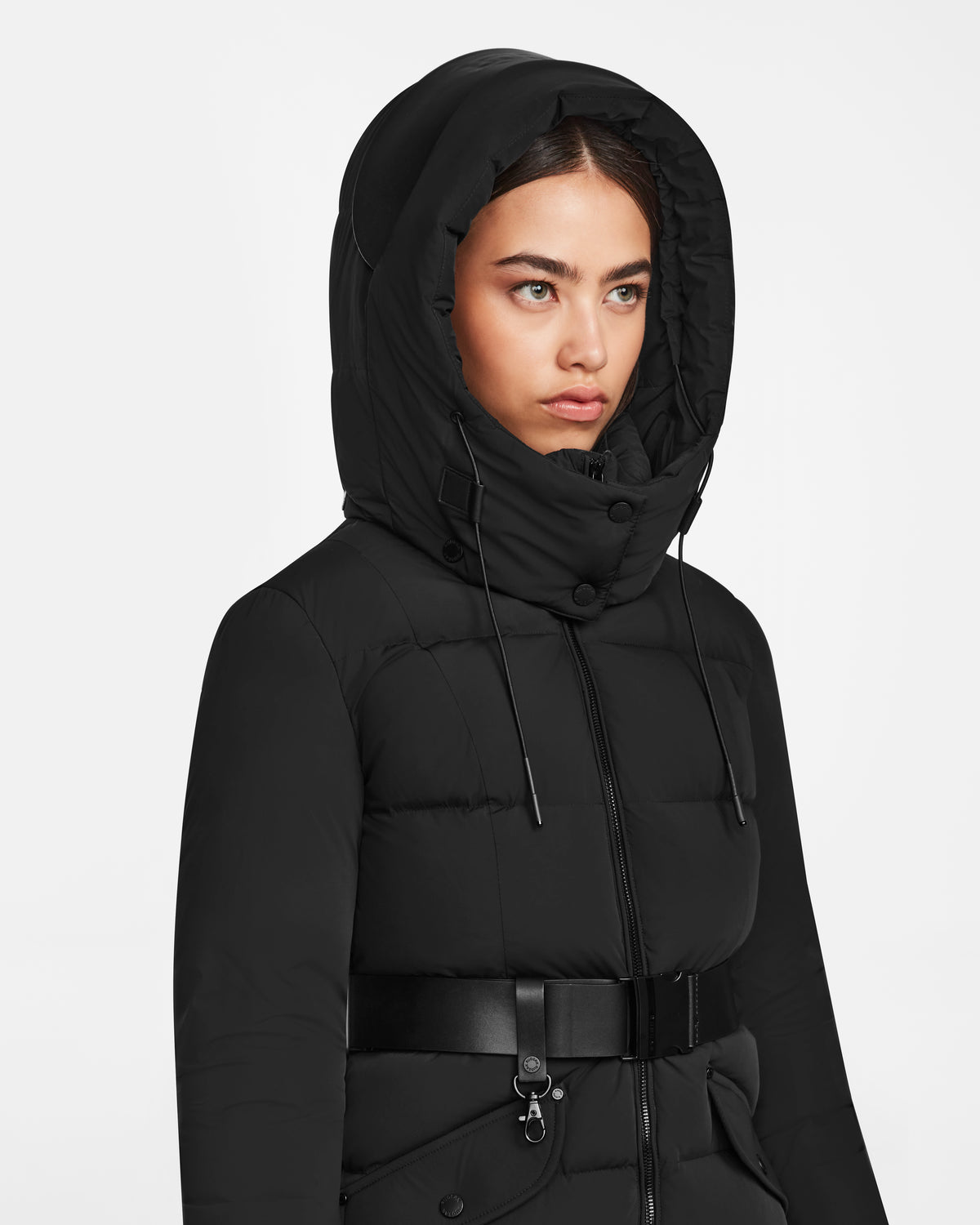A woman wears the Rudsak CAT - 8124584 Black mid-length quilted down puffer with a large hood and belted waist, gazing off to the side with a neutral expression against a plain white background.