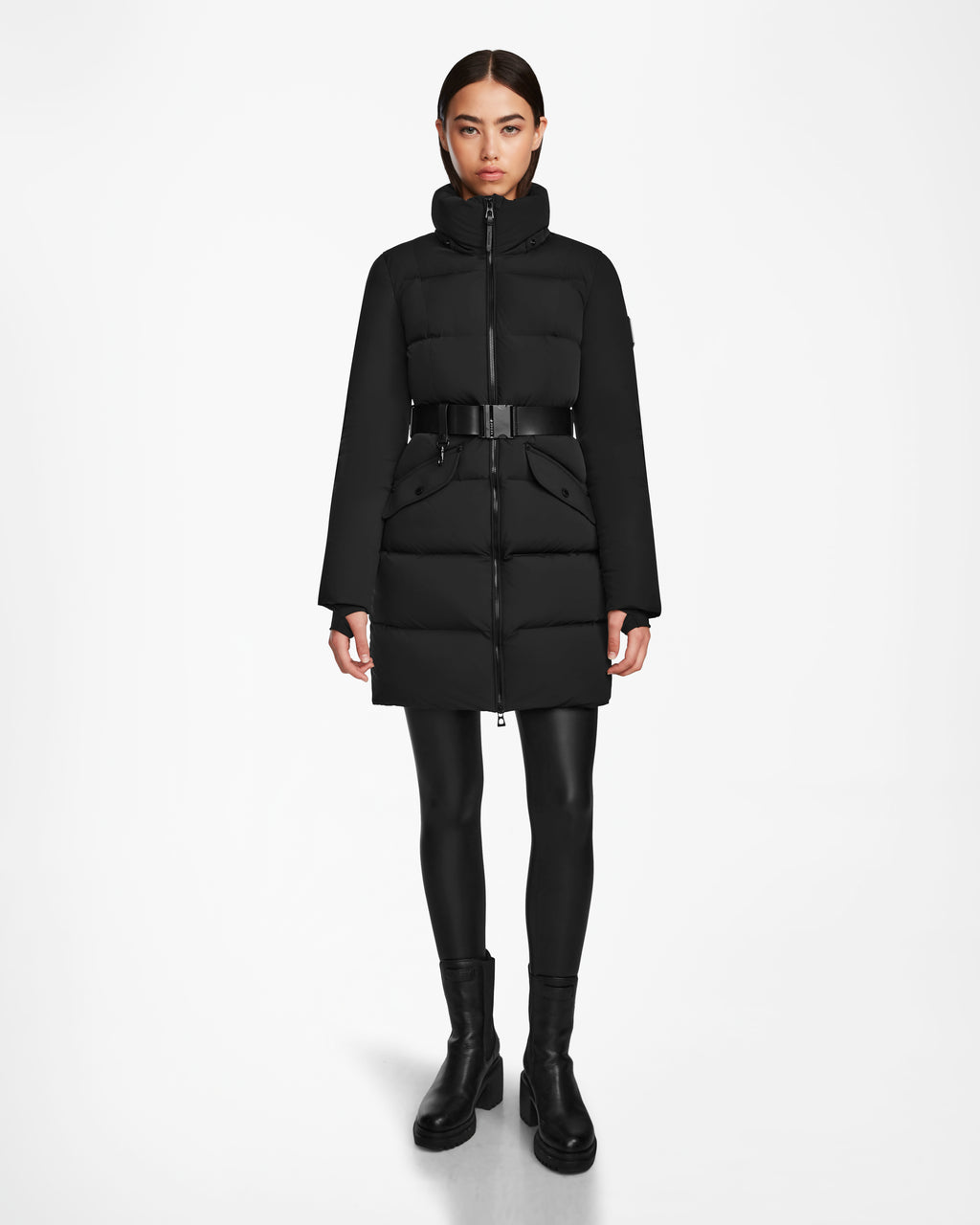 A woman faces forward against a white background, wearing the Rudsak CAT - 8124584 Black mid-length belted puffer coat, styled with black leggings and ankle boots.
