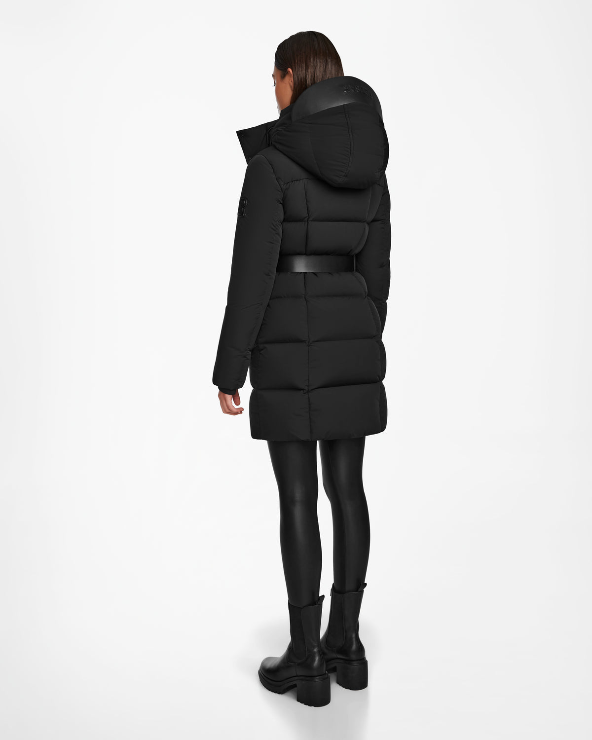 A woman wearing the Rudsak CAT - 8124584 Black mid-length puffer coat stands facing away, with black leggings and boots, against a plain white background.