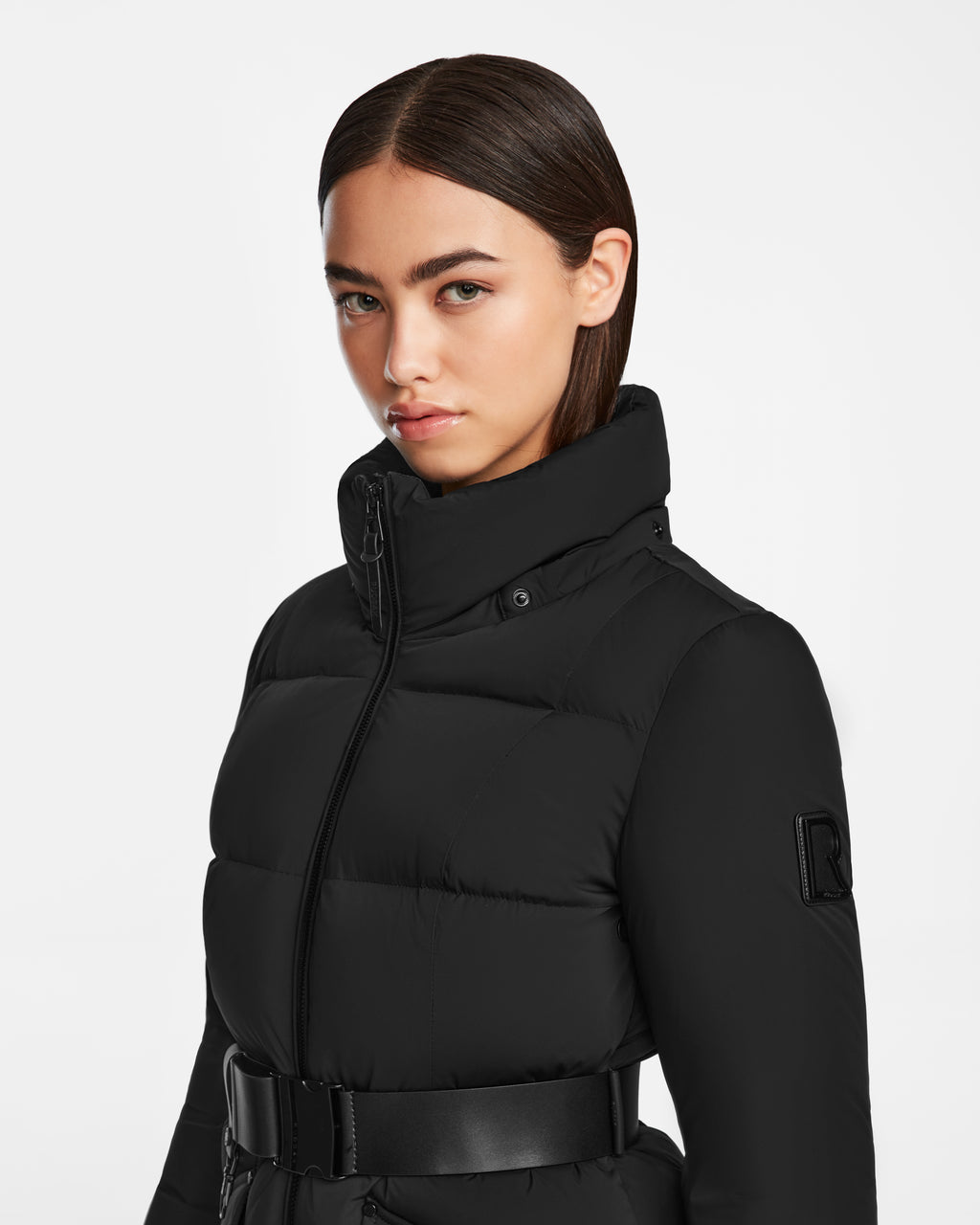 A woman with straight brown hair, parted in the middle, models the Rudsak CAT - 8124584 Black quilted down puffer with a high collar and belted waist. She faces slightly sideways against a plain white background.