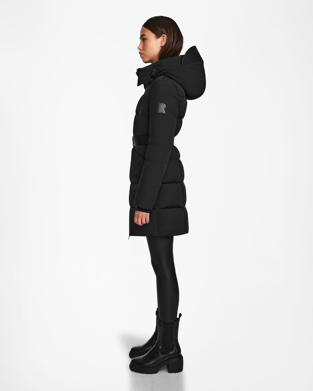 A woman in profile wears the Rudsak CAT - 8124584 Black womens quilted puffer coat with hood and RDS-certified down, paired with black leggings and ankle boots. The mid-length jacket is shown against a plain white background.