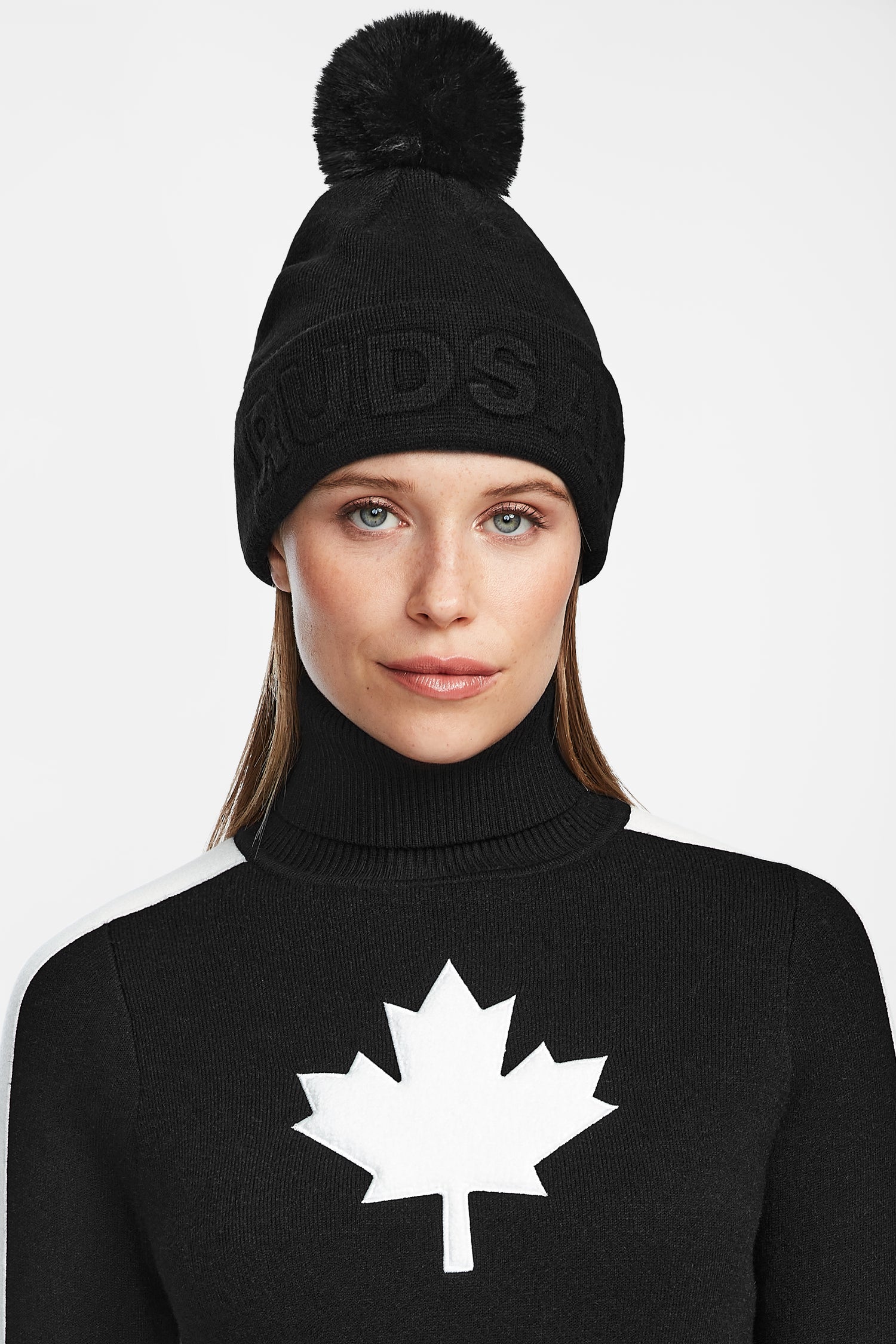 A woman models the Rudsak CERRY - 9423508 Black wool blend winter hat with removable pom pom and a black turtleneck sweater featuring a large white maple leaf, set against a plain white background.