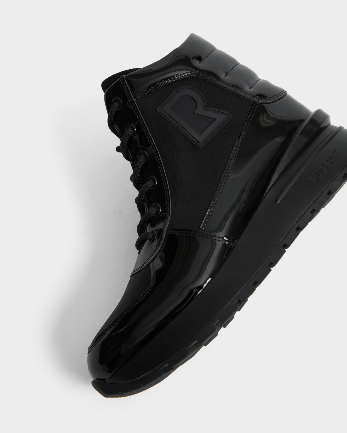 The Rudsak CHELSA - L224505 Black is a women’s black high-top sneaker boot with glossy and matte finishes, black laces, a bold “R” side logo, and a waterproof zipper. Shown angled to reveal the side and sole against a white background.