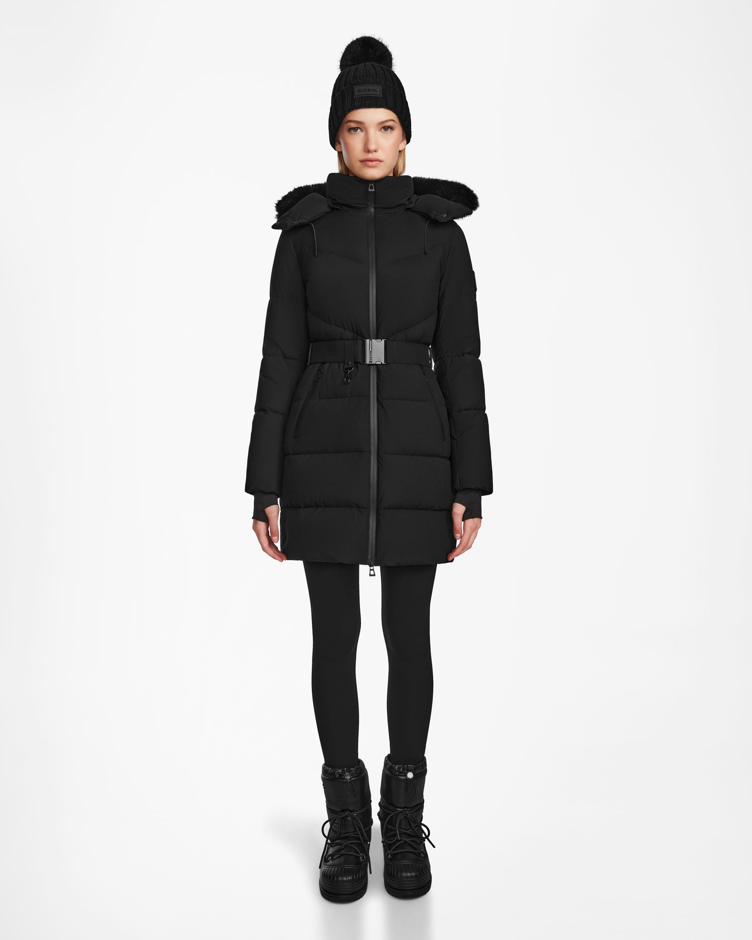 A person stands facing forward wearing the Rudsak CHRISTEL M-M - L124505 Black quilted winter coat with a belt, paired with black leggings, boots, and a pom-pom knit hat, against a plain white background.