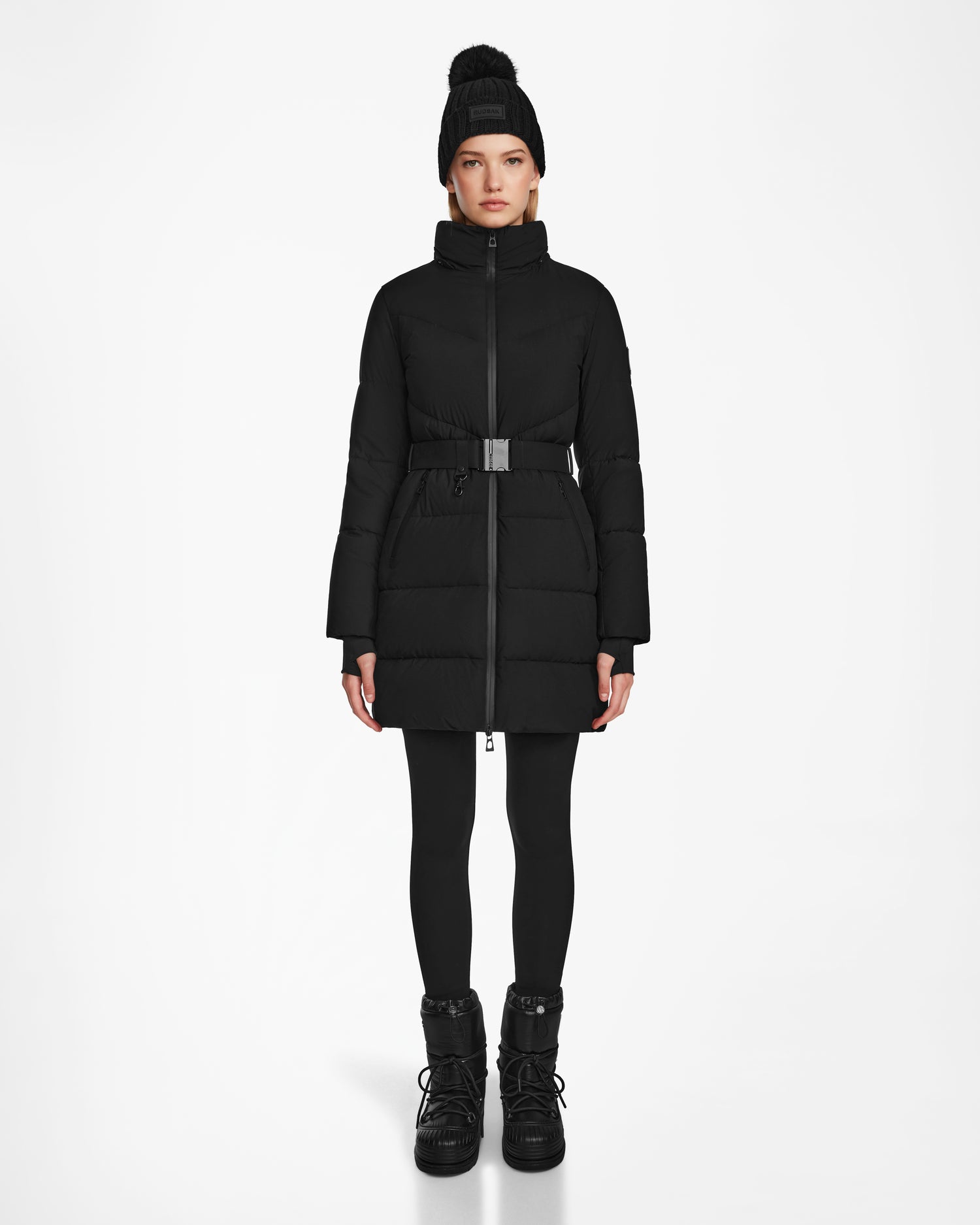 A person wearing the Rudsak CHRISTEL M-M - L124505 Black down puffer with a belt, paired with black leggings, winter boots, and a black pom-pom knit hat, stands against a plain white background.