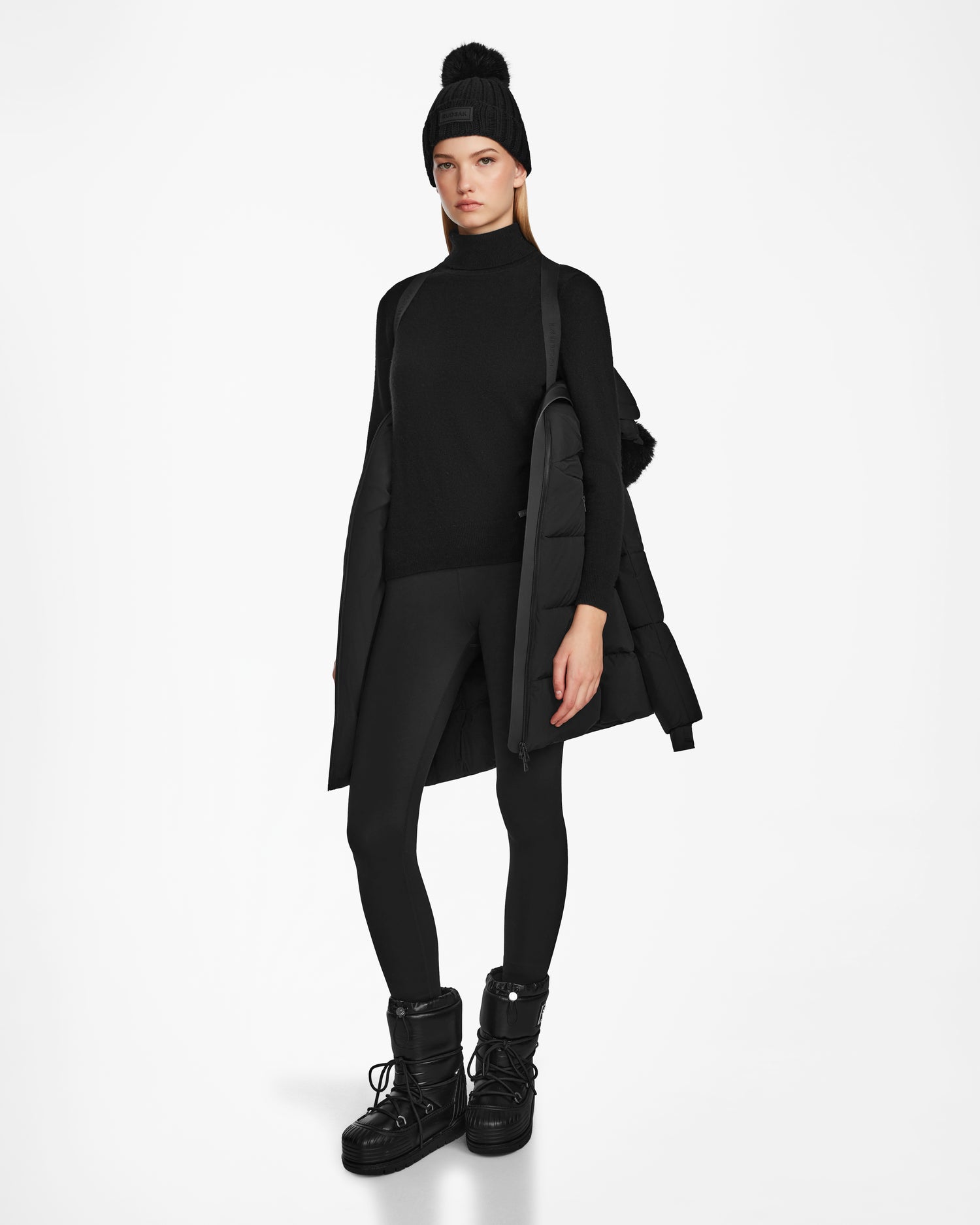 A person with a neutral expression stands against a white background wearing the Rudsak CHRISTEL M-M - L124505 Black quilted winter coat draped over their shoulders. They’re also dressed in a black beanie, turtleneck, leggings, and winter boots.