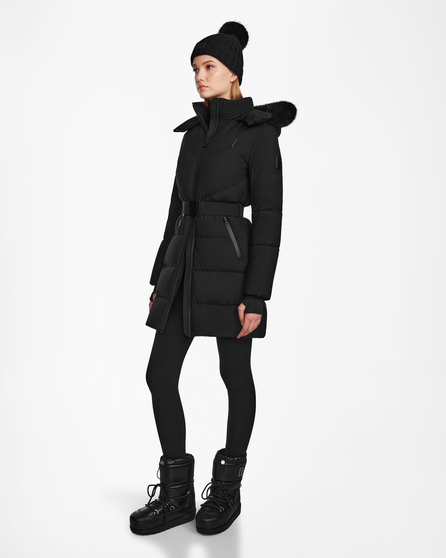 A person wearing the Rudsak CHRISTEL M-M - L124505 Black quilted winter coat with a fur-trimmed hood stands against a plain white background, styled with black leggings, black boots, and a black knit beanie.