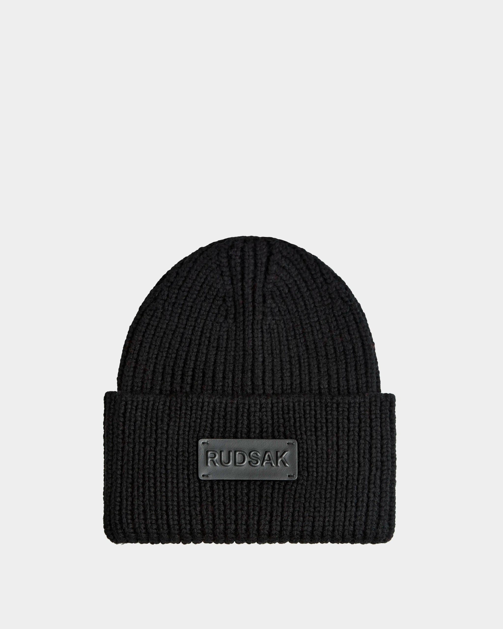 The COLIN - 9424518 Black by Rudsak is a black unisex ribbed knit hat with a wide folded brim and a rectangular RUDSAK patch on the front, displayed on a plain light background.
