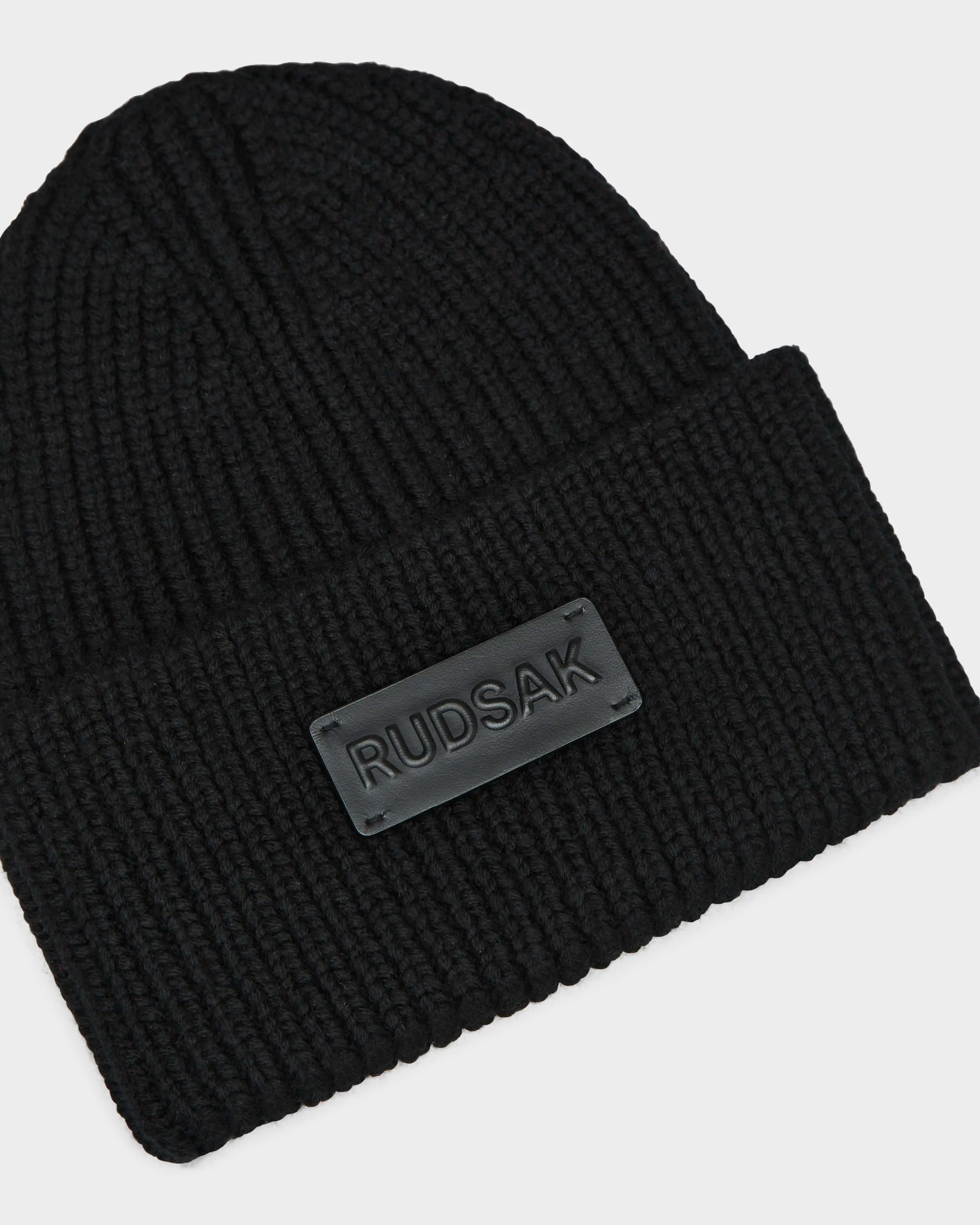 The COLIN - 9424518 Black by Rudsak is a black unisex beanie with a folded brim, featuring a rectangular black patch label embossed with RUDSAK on the front.