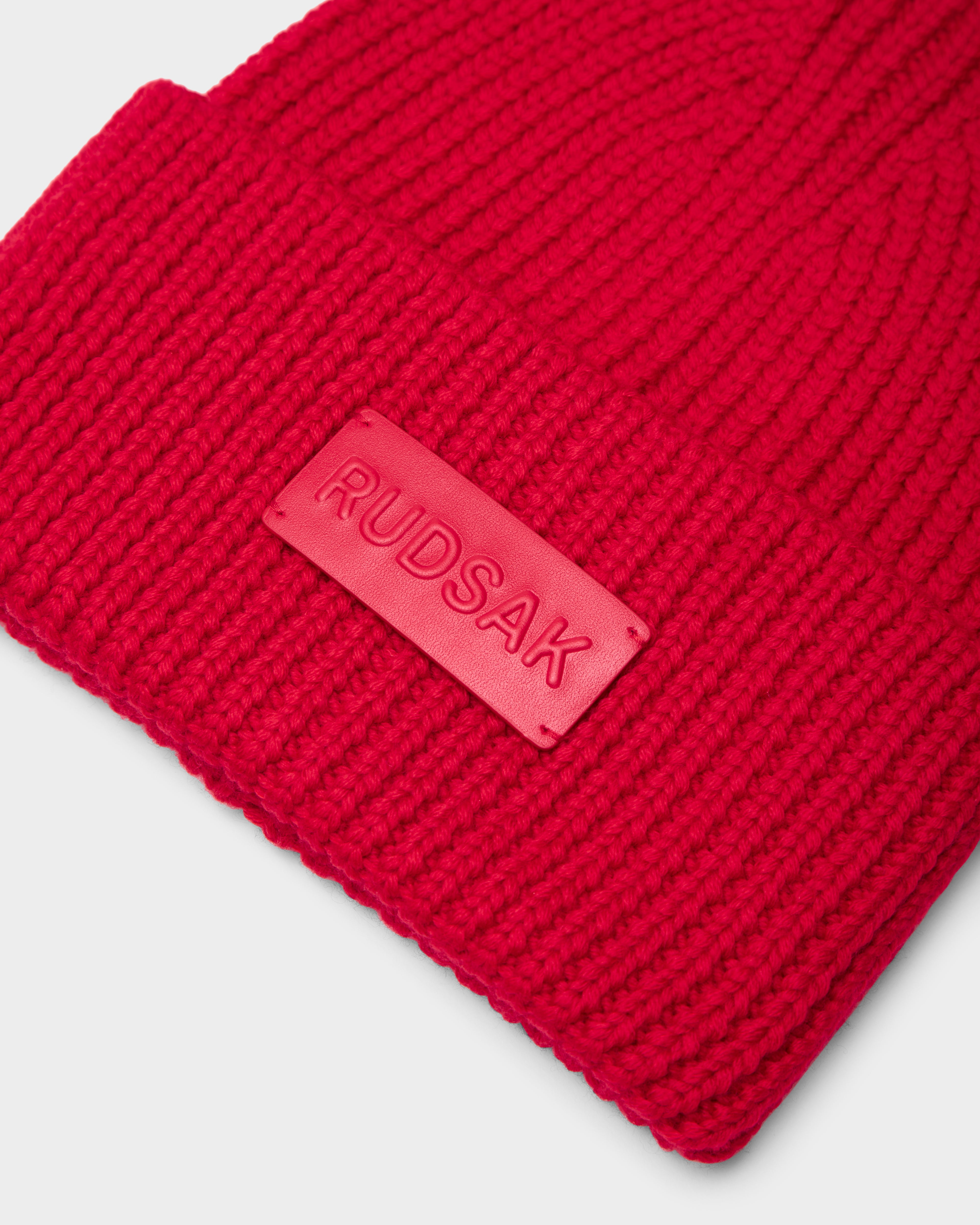 A close-up of the Rudsak COLIN - 9424518 Flame Red unisex beanie with a wide folded cuff and a rectangular rubber RUDSAK patch, set against a plain light gray background.