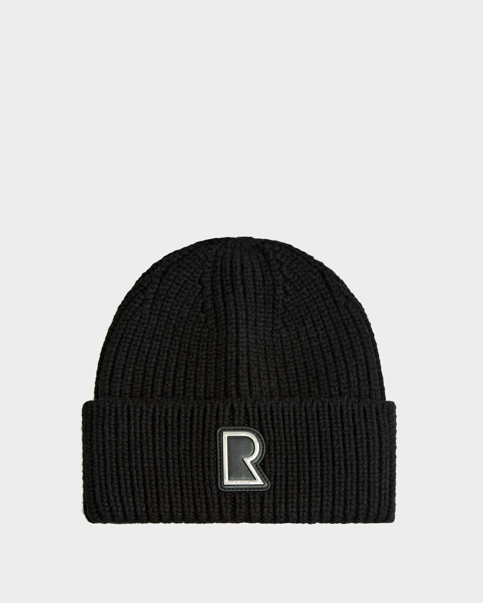 The Rudsak CYBELLE - 9424504 Black/Silver is a black ribbed knit winter beanie with a folded cuff and a large front patch featuring an outlined white R, shown on a plain light gray background.