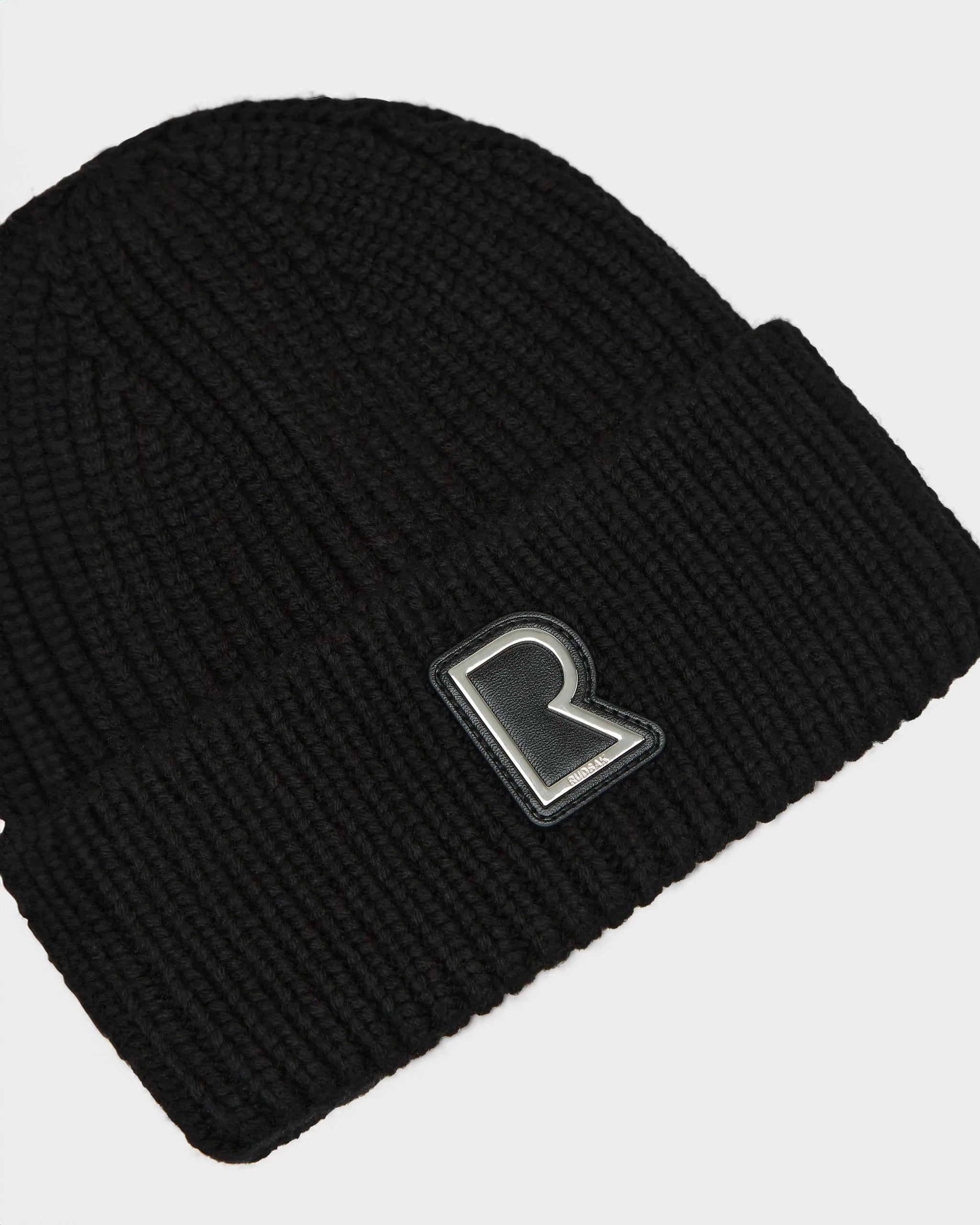 The CYBELLE - 9424504 Black/Silver by Rudsak is a unisex black winter beanie with a folded cuff and metallic letter R patch on the front, shown against a plain white background.