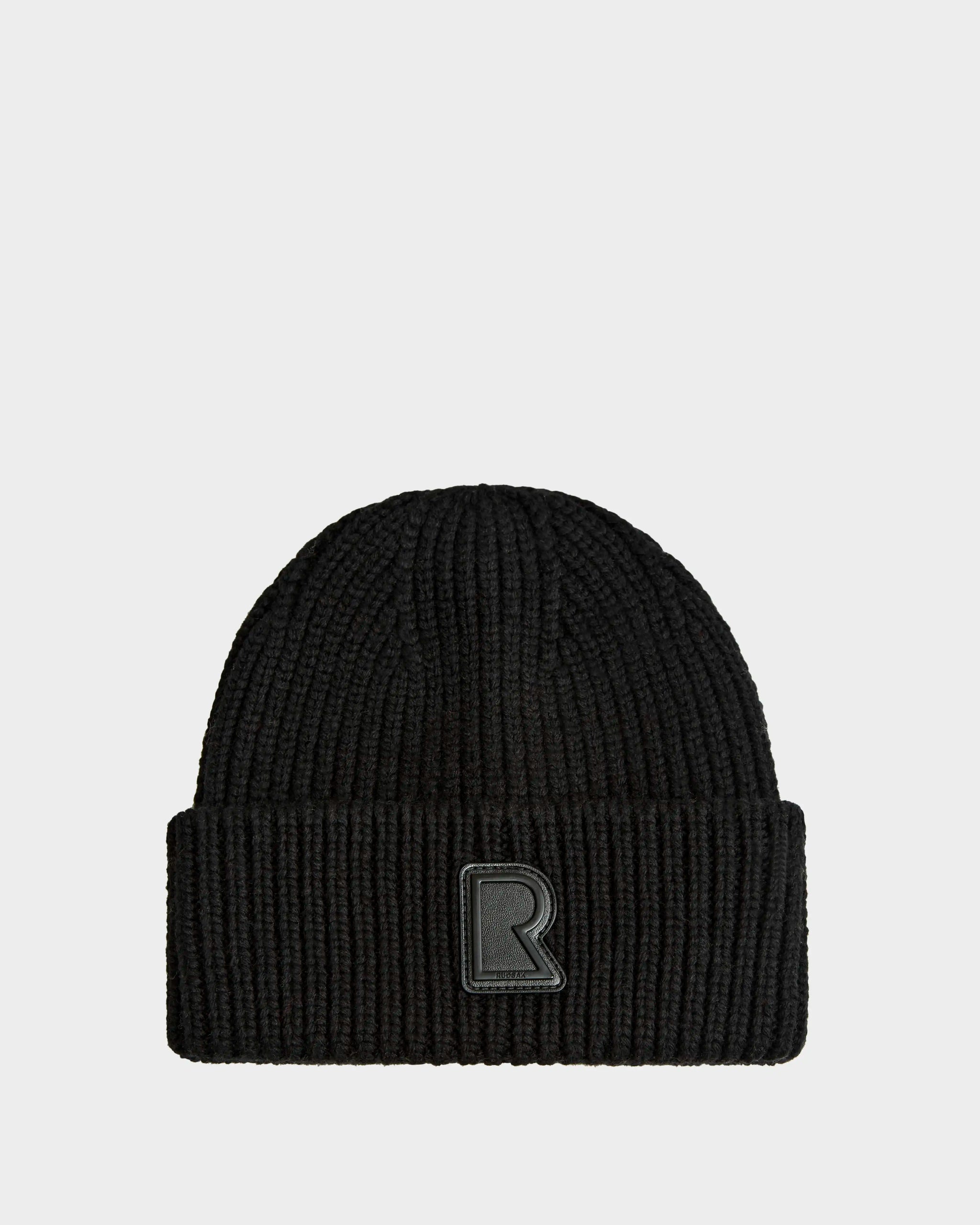 The Rudsak CYBELLE - 9424504 Black is a unisex black ribbed knit wool beanie with a folded brim and raised black R patch on the front, shown on a plain light gray background.