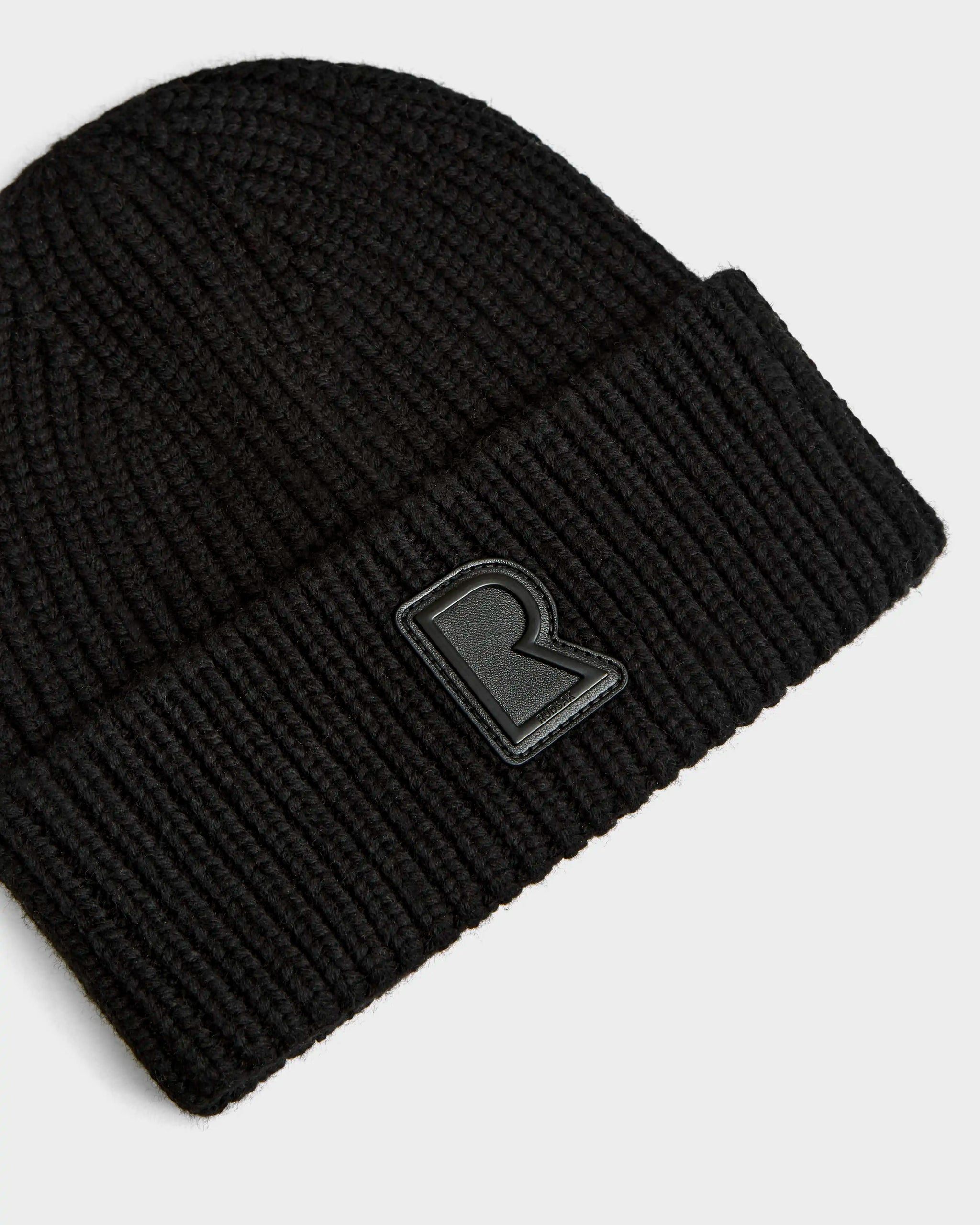 A close-up of the Rudsak CYBELLE - 9424504 Black unisex ribbed knit hat with a folded brim, featuring a raised black R patch on the front, displayed against a plain white background.