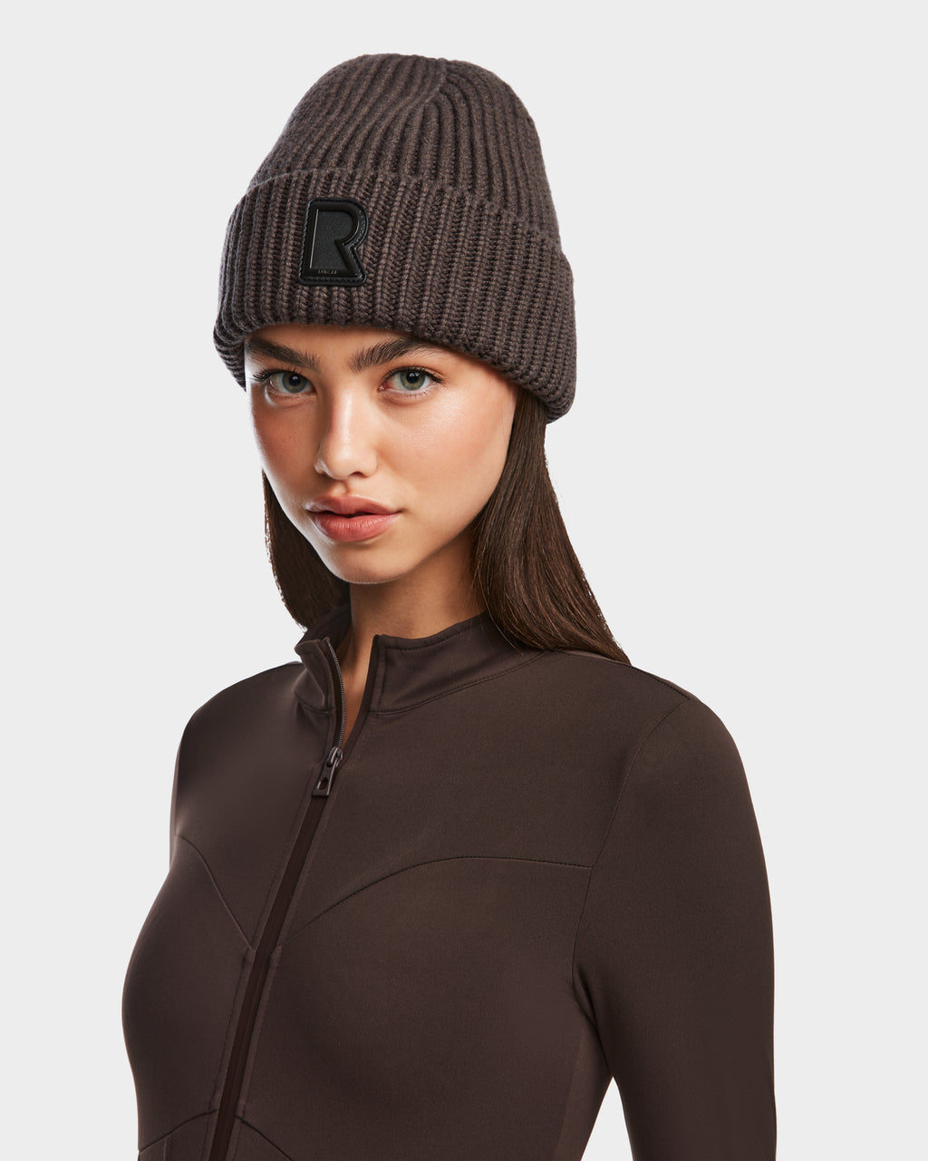 A woman wears the Rudsak CYBELLE - 9424504 Espresso ribbed brown wool blend beanie with a black R patch, paired with a fitted brown zip-up top, standing against a plain light background.