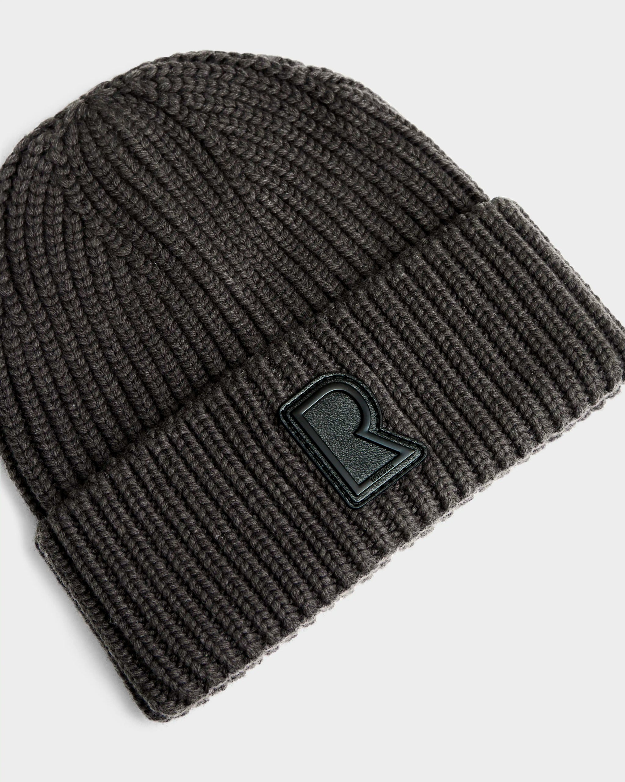The Rudsak CYBELLE - 9424504 Grey is a dark gray unisex knit hat with a wide cuffed brim and a black patch featuring a raised letter R on the front, shown here against a light background.