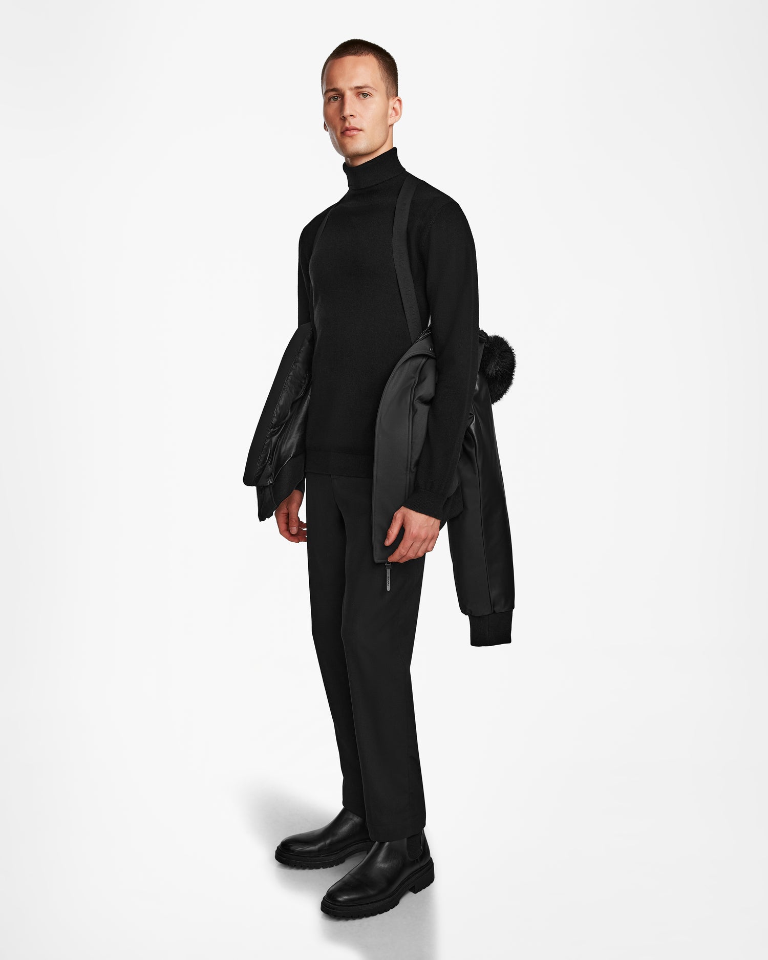 A man stands against a white background wearing black clothes and holds the Rudsak DALLAS R - M123501 BLACK men's quilted down bomber with recycled leather sleeves over his shoulder, looking straight ahead with a neutral expression.