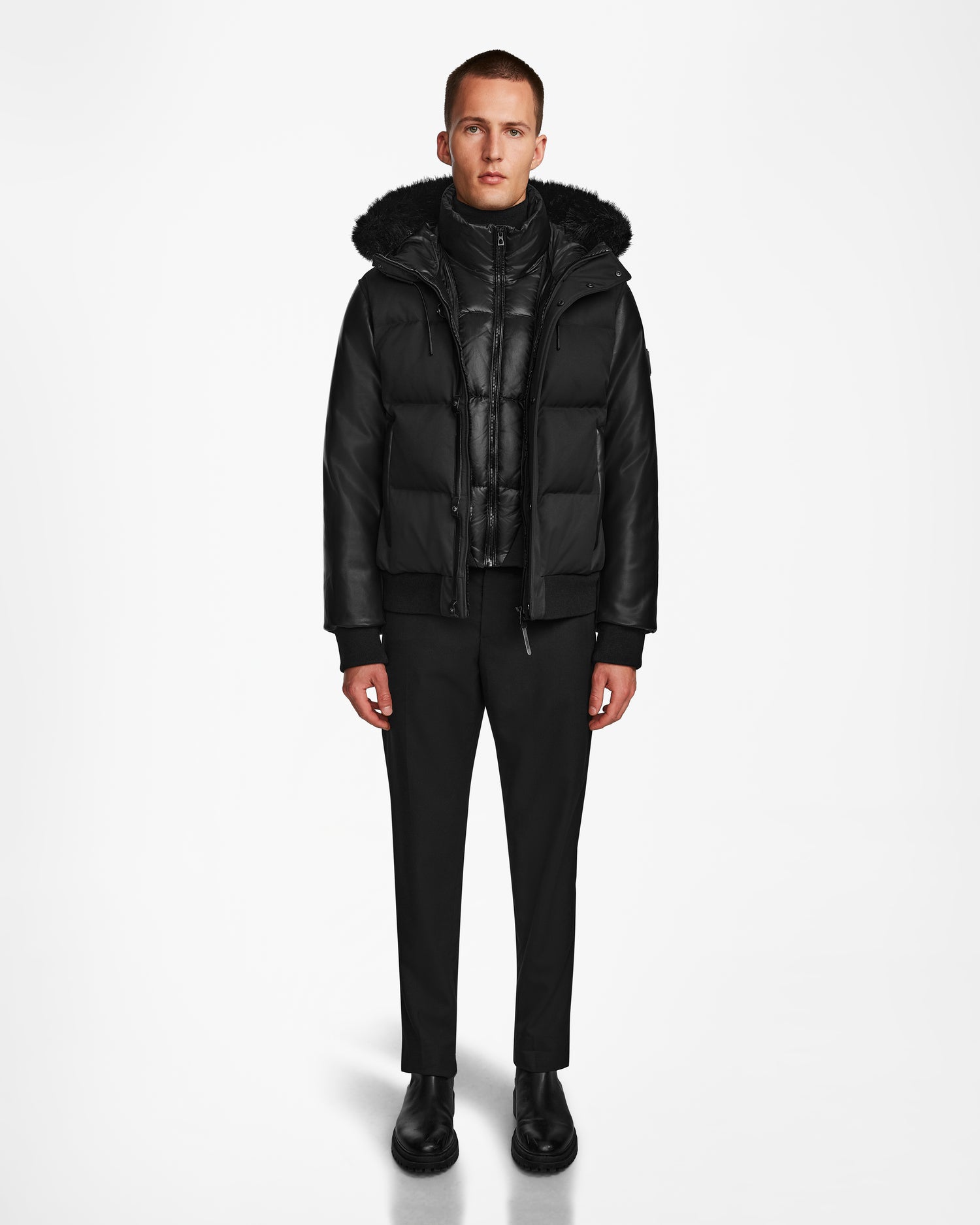 A person stands facing forward against a plain white background, wearing the Rudsak DALLAS R - M123501 BLACK— a weather-resistant hooded puffer jacket with recycled leather sleeves—paired with black pants and shoes.