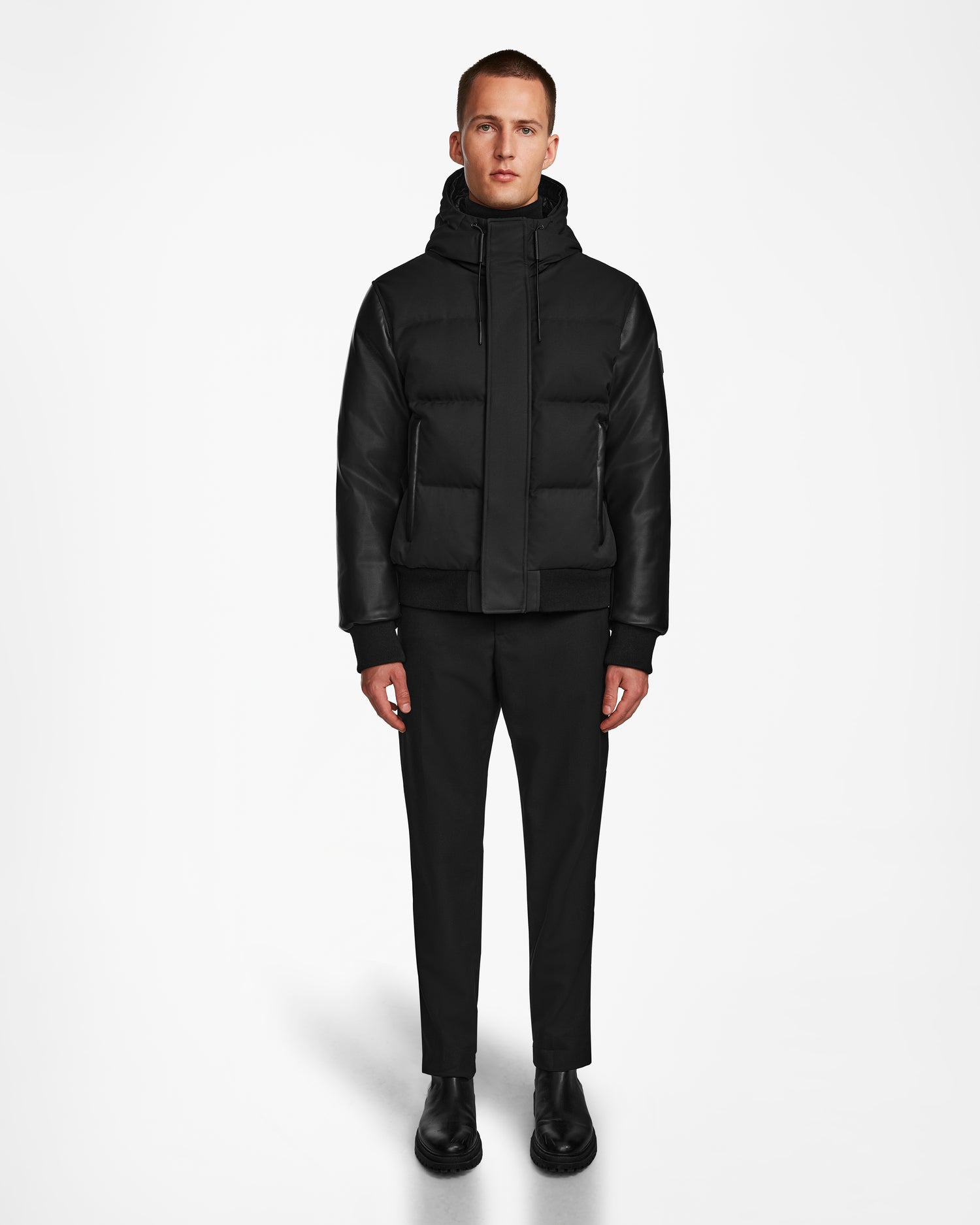 A person faces forward wearing the Rudsak DALLAS R - M123501 BLACK men's quilted down bomber with weather-resistant fabric and recycled leather sleeves, paired with black pants and shoes, on a plain white background.