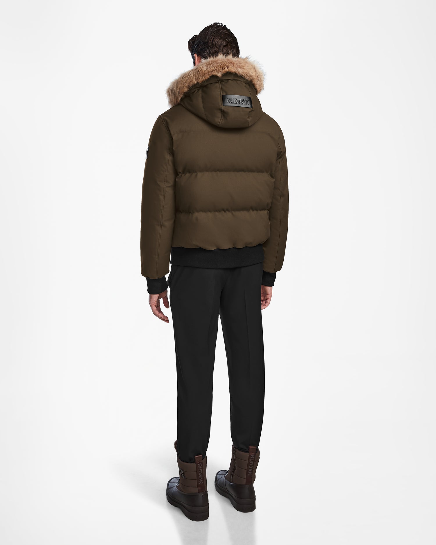 A person faces away, wearing the Rudsak DALLAS - M122501 Army weather-resistant puffer with a fur-lined hood, black pants, and brown boots against a plain white background.