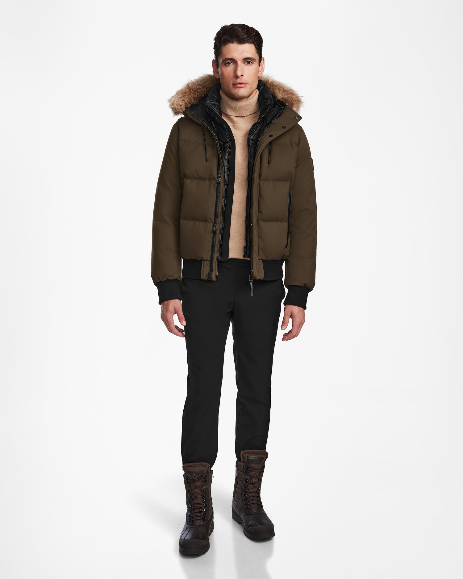A man faces forward against a white background, wearing the Rudsak DALLAS - M122501 Army weather-resistant puffer jacket with fur-trimmed hood, plus a beige sweater, black pants, and brown lace-up boots.