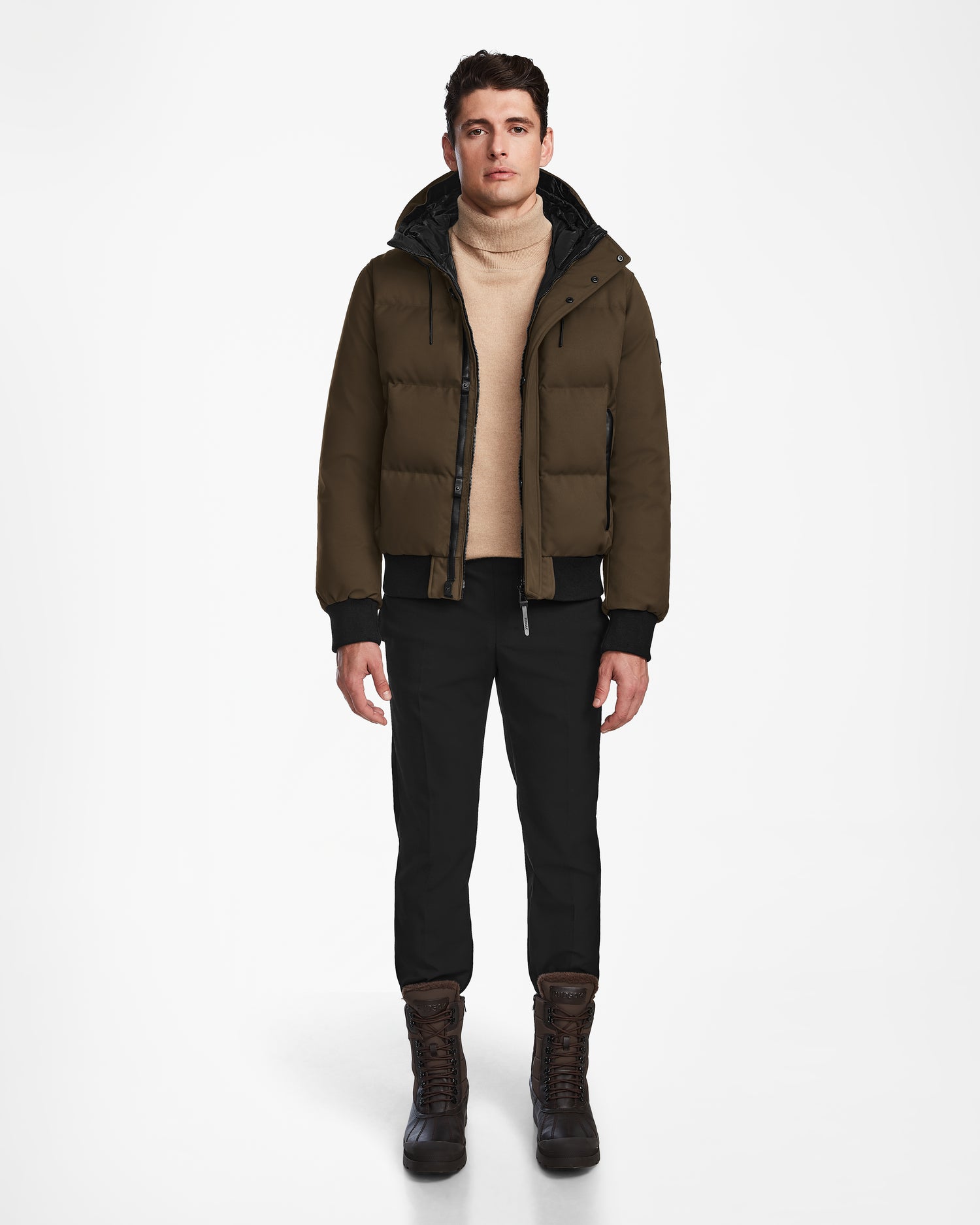 A man stands facing forward against a plain white background, wearing the Rudsak DALLAS - M122501 Army weather-resistant puffer jacket, a beige turtleneck sweater, black pants, and brown winter boots.
