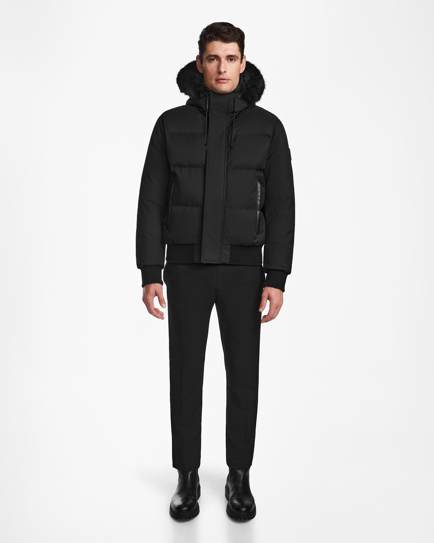 A man stands facing forward in a Rudsak DALLAS - M122501 black down bomber puffer coat with a fur-lined hood, paired with black pants and boots, set against a plain white background.