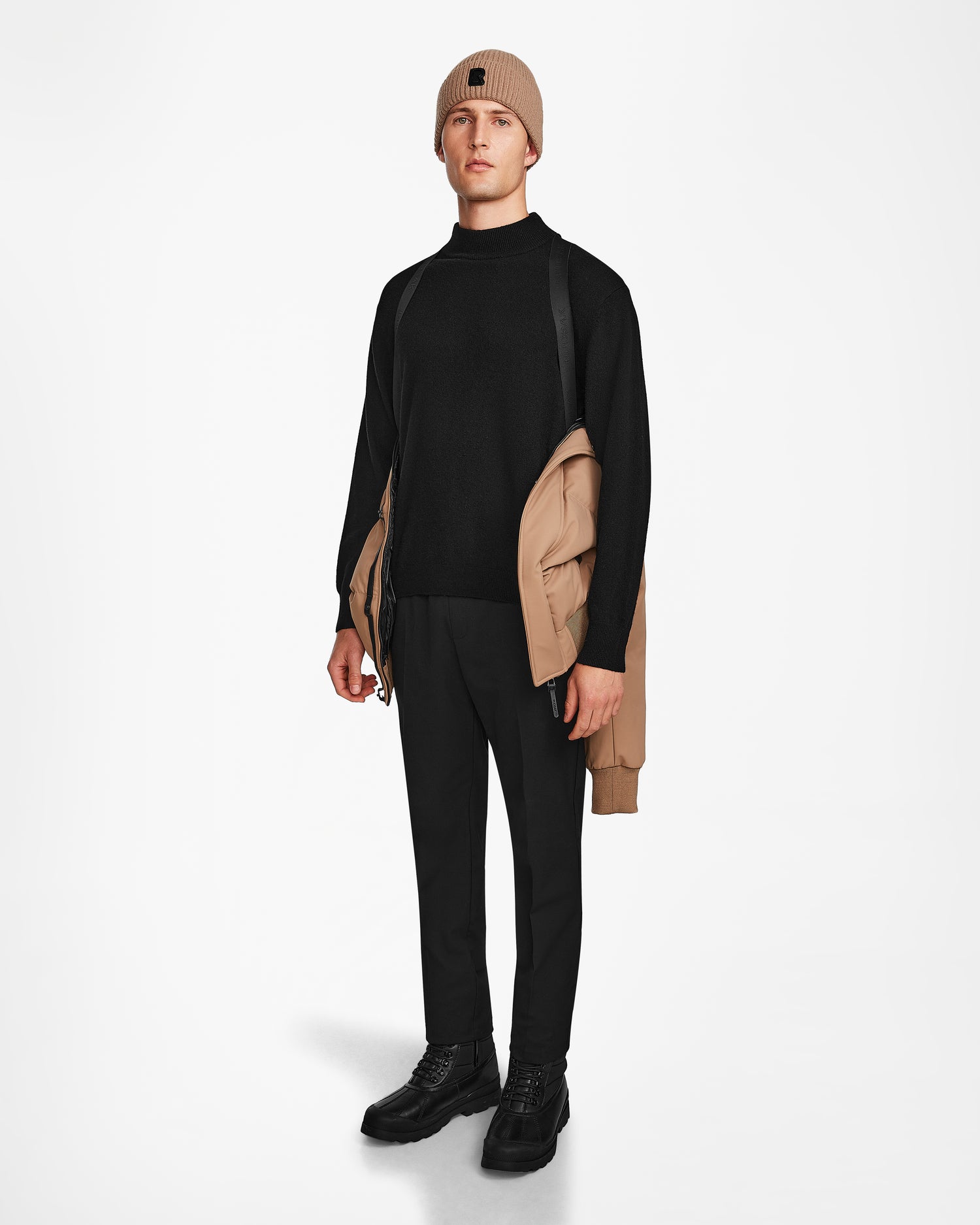 A person stands facing forward, holding the Rudsak DALLAS - M122501 Sand men's quilted down bomber over one shoulder, dressed in a beige beanie, black sweater and pants, and black shoes against a plain white background.