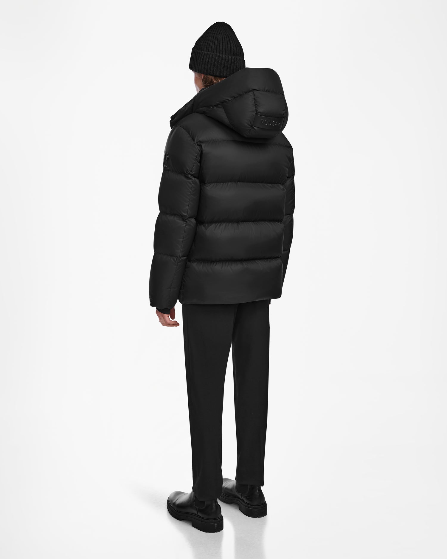 A person faces away, styled in Rudsak’s DANIEL M - M124510 Black men’s hooded down puffer with a black beanie, black pants, and black shoes against a plain white background.