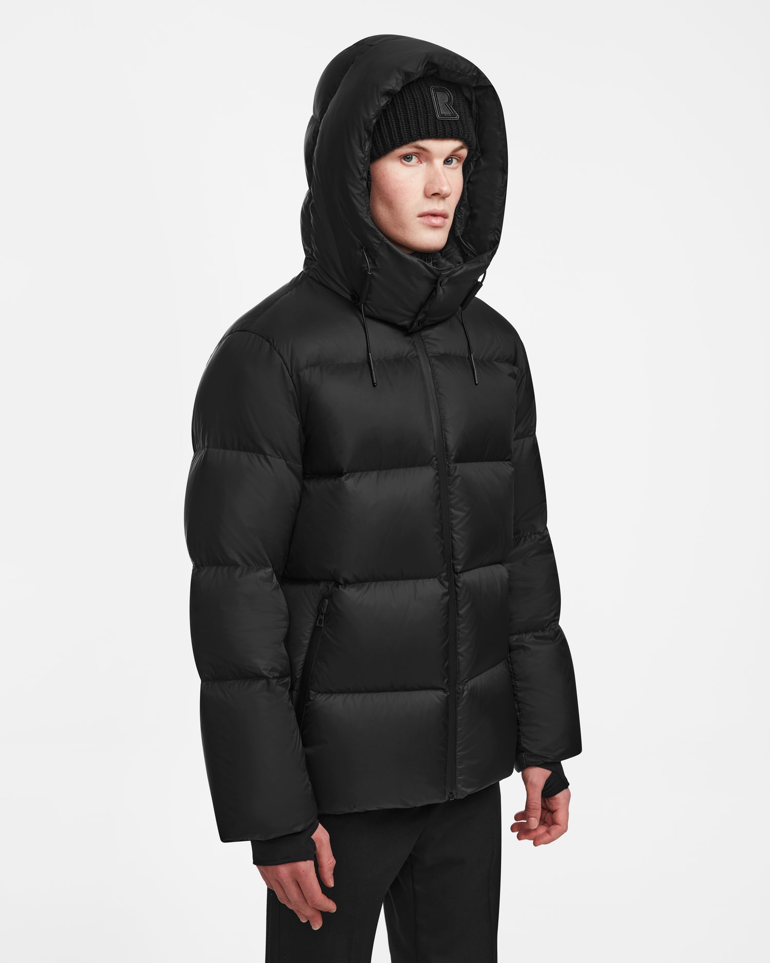 A person wearing the Rudsak DANIEL M - M124510 Black mens hooded down puffer jacket and a black beanie stands against a plain white background.
