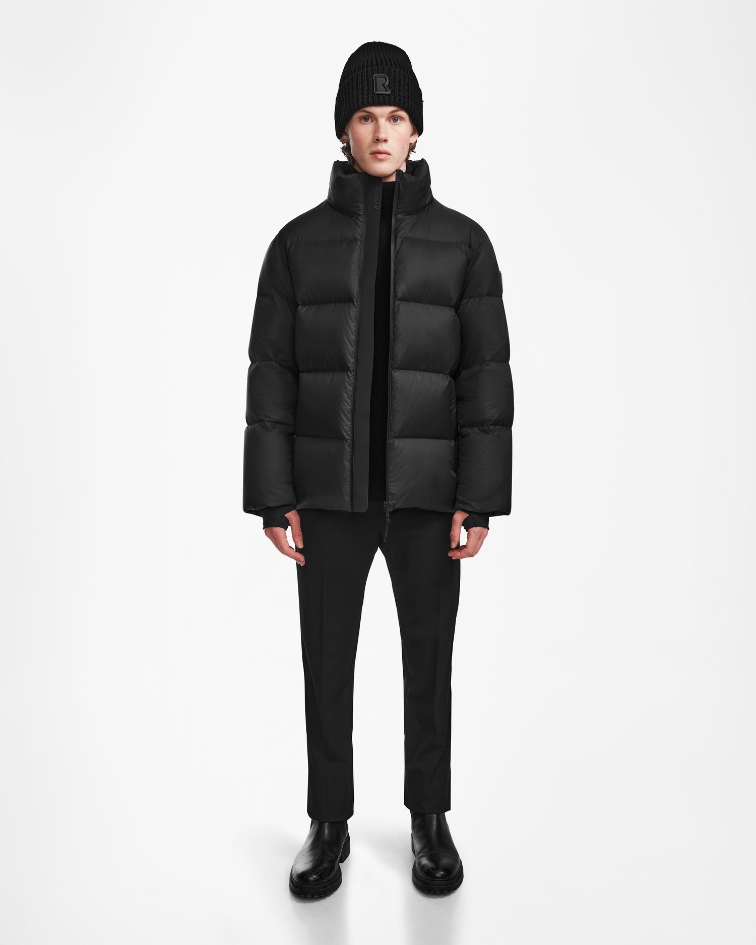 A person stands facing forward against a plain white background, wearing the Rudsak DANIEL M - M124510 Black weather-resistant puffer jacket with black pants, boots, and a beanie.