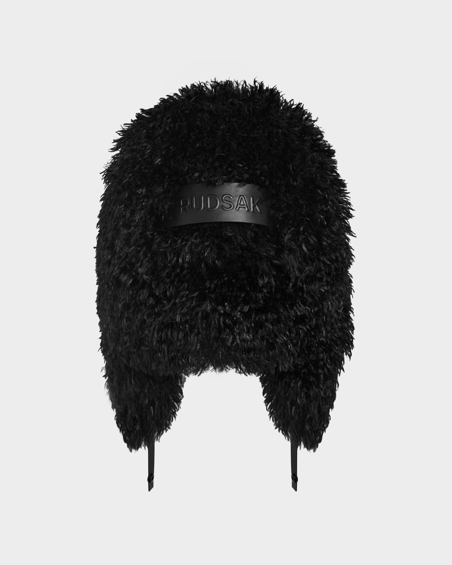 The Rudsak DARBY M - 9424510 Black is a unisex aviator-style faux Mongolian fur trapper hat with ear flaps, a leather front patch, and a fluffy black finish—ideal as a stylish winter accessory. Photographed on a white background.