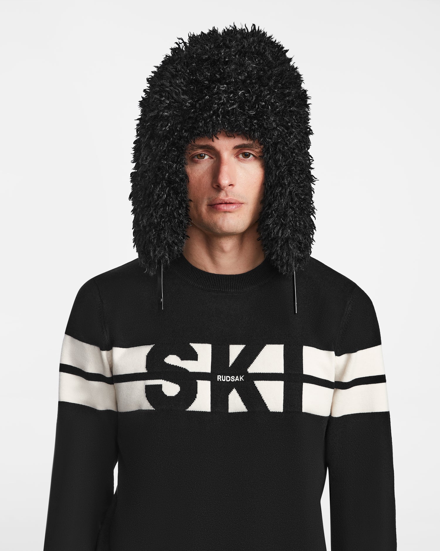 A man wears the Rudsak DARBY M - 9424510 Black unisex aviator hat with faux Mongolian fur ear flaps and a black sweater with white stripes and SKI on the front, against a plain white background.