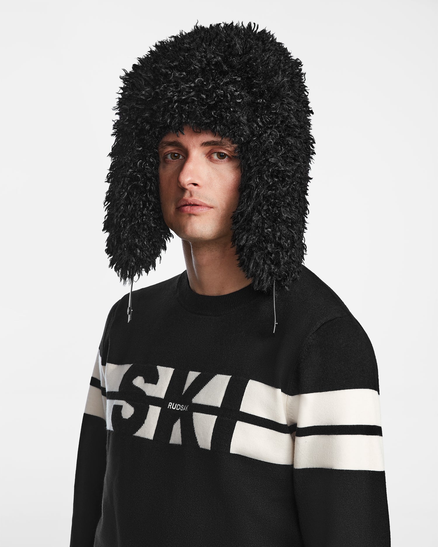 Wearing the Rudsak DARBY M - 9424510 Black unisex aviator-style hat with faux Mongolian fur and ear flaps, and a black sweater with bold white stripes and "SKI" on the front, a person stands against a plain white background.